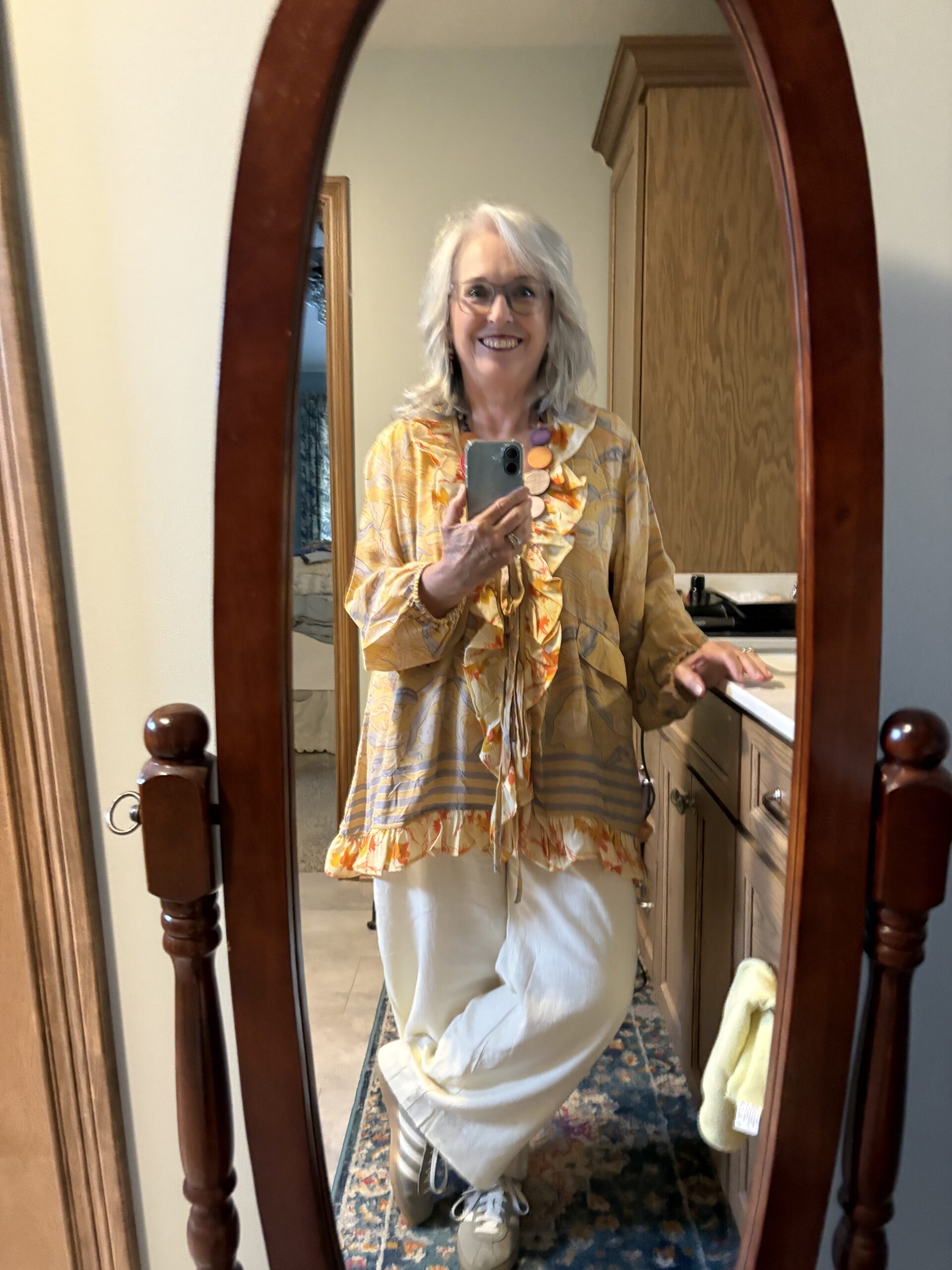 Woman taking a mirror selfie in a wooden-framed oval mirror, wearing a yellow patterned blouse and light-colored pants, smiling at the camera.