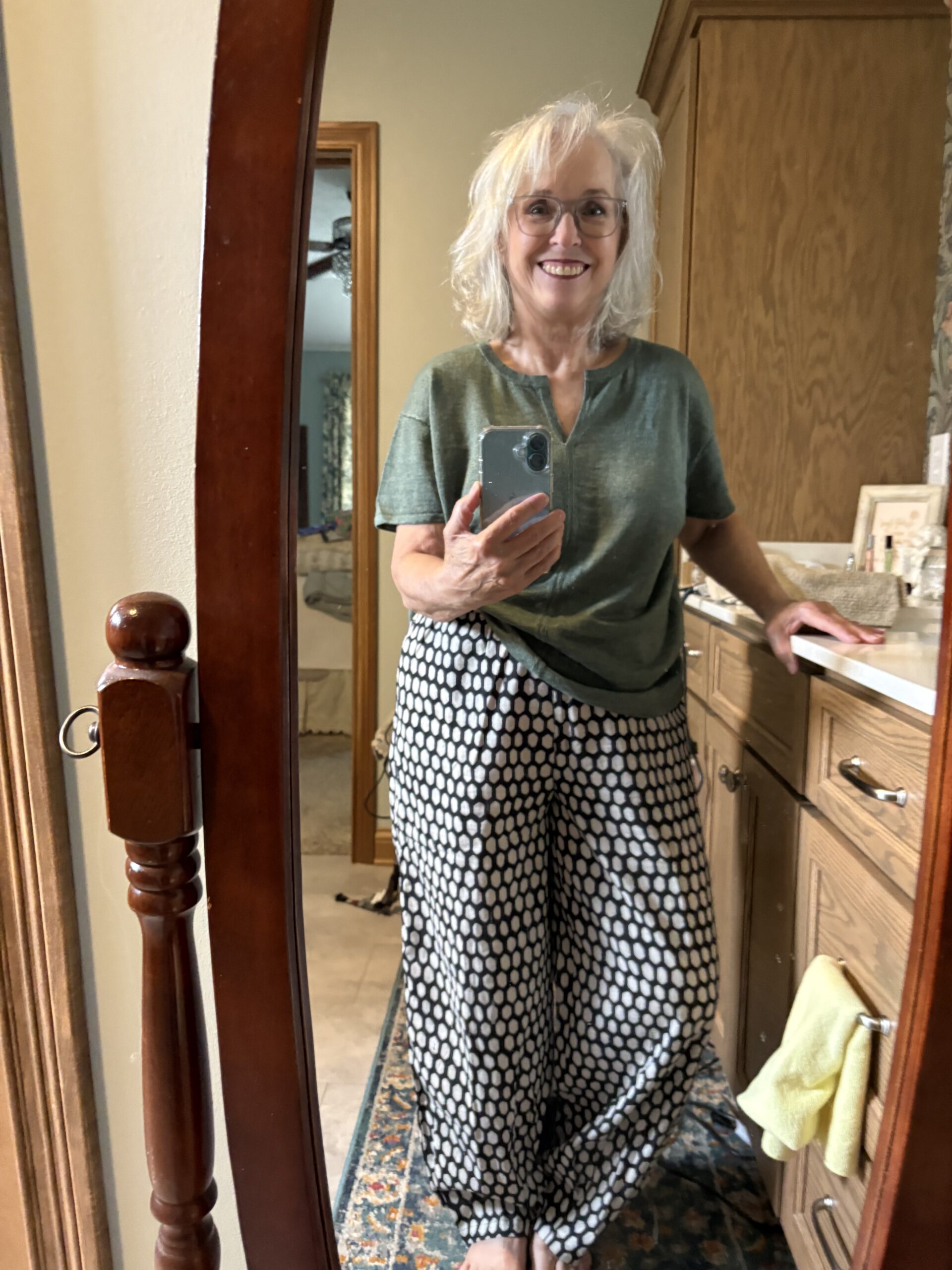 Older woman with gray hair and glasses taking a mirror selfie in a bathroom, wearing a green top and a black-and-white polka dot skirt.