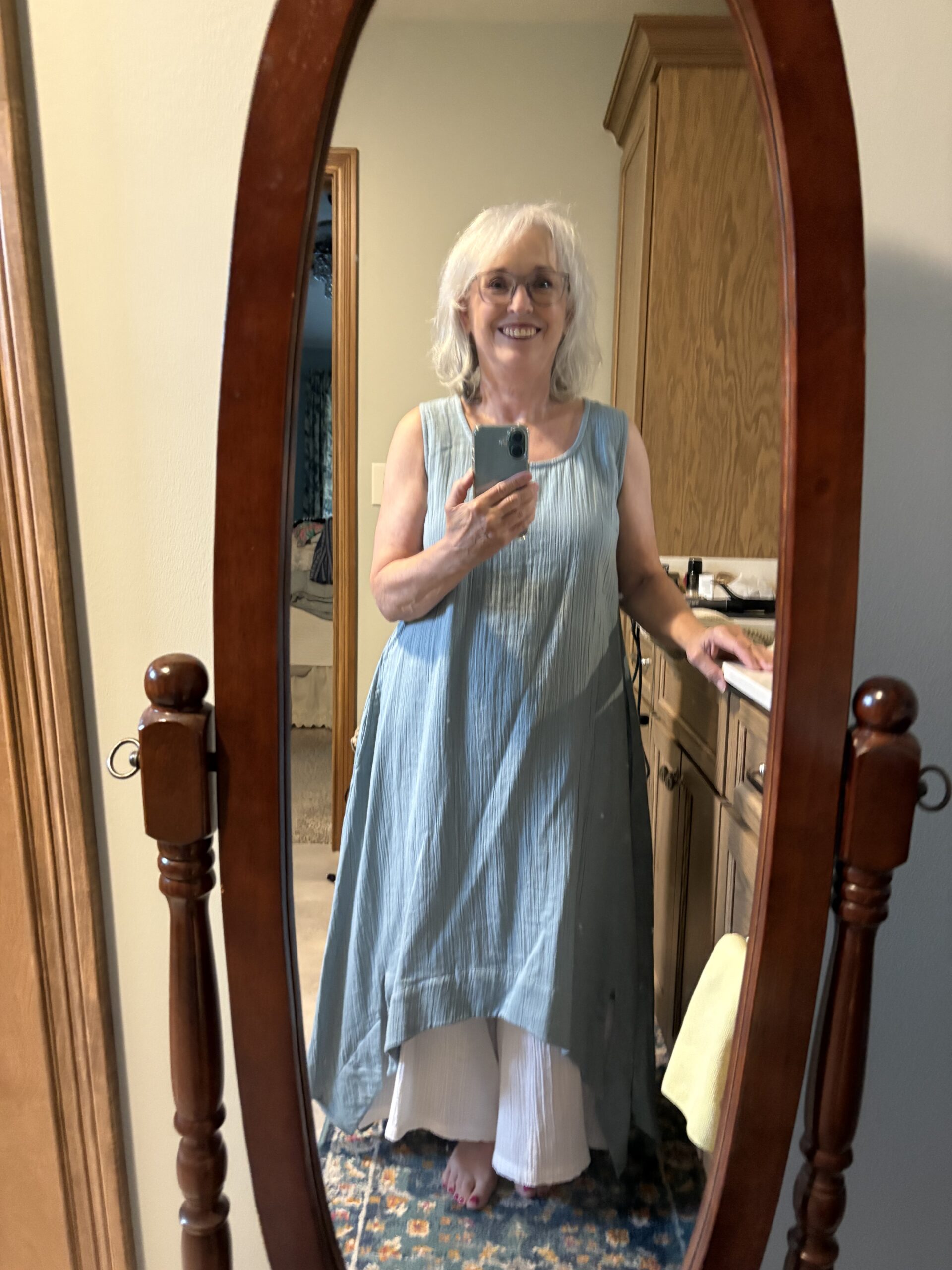 Smiling older woman in a light blue sleeveless dress taking a mirror selfie in a bedroom, holding a smartphone.