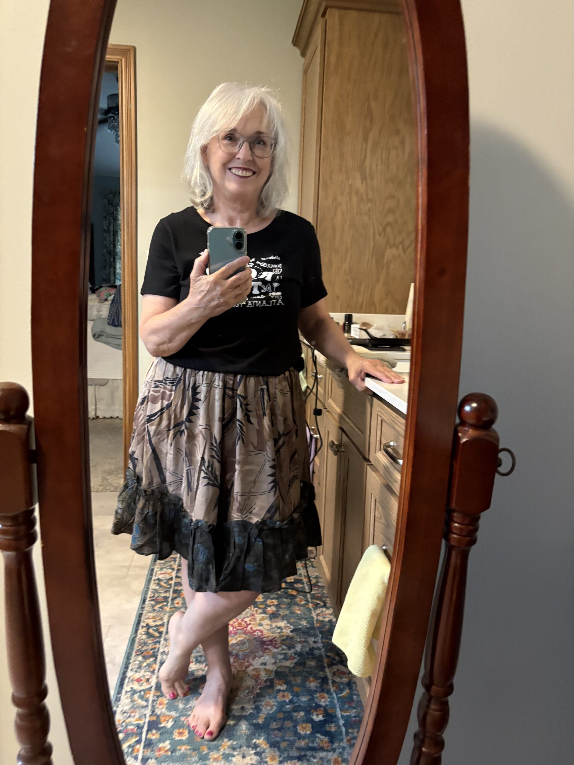 Smiling older woman with white hair and glasses taking a mirror selfie in a bedroom, wearing a black top and patterned skirt.