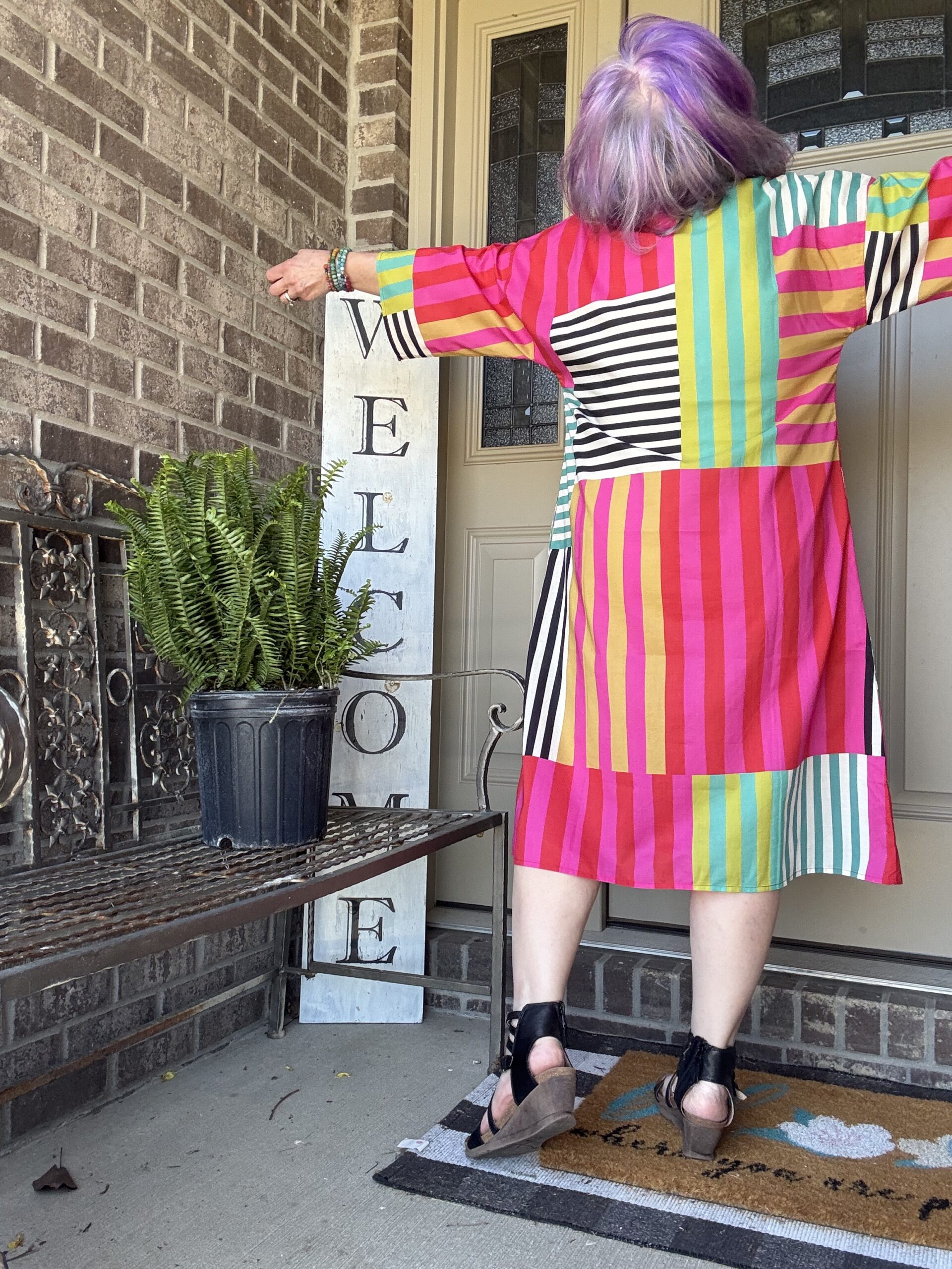 Person with purple hair wearing a bright, multi-colored striped dress stands on a porch with arms outstretched, facing away from the camera.