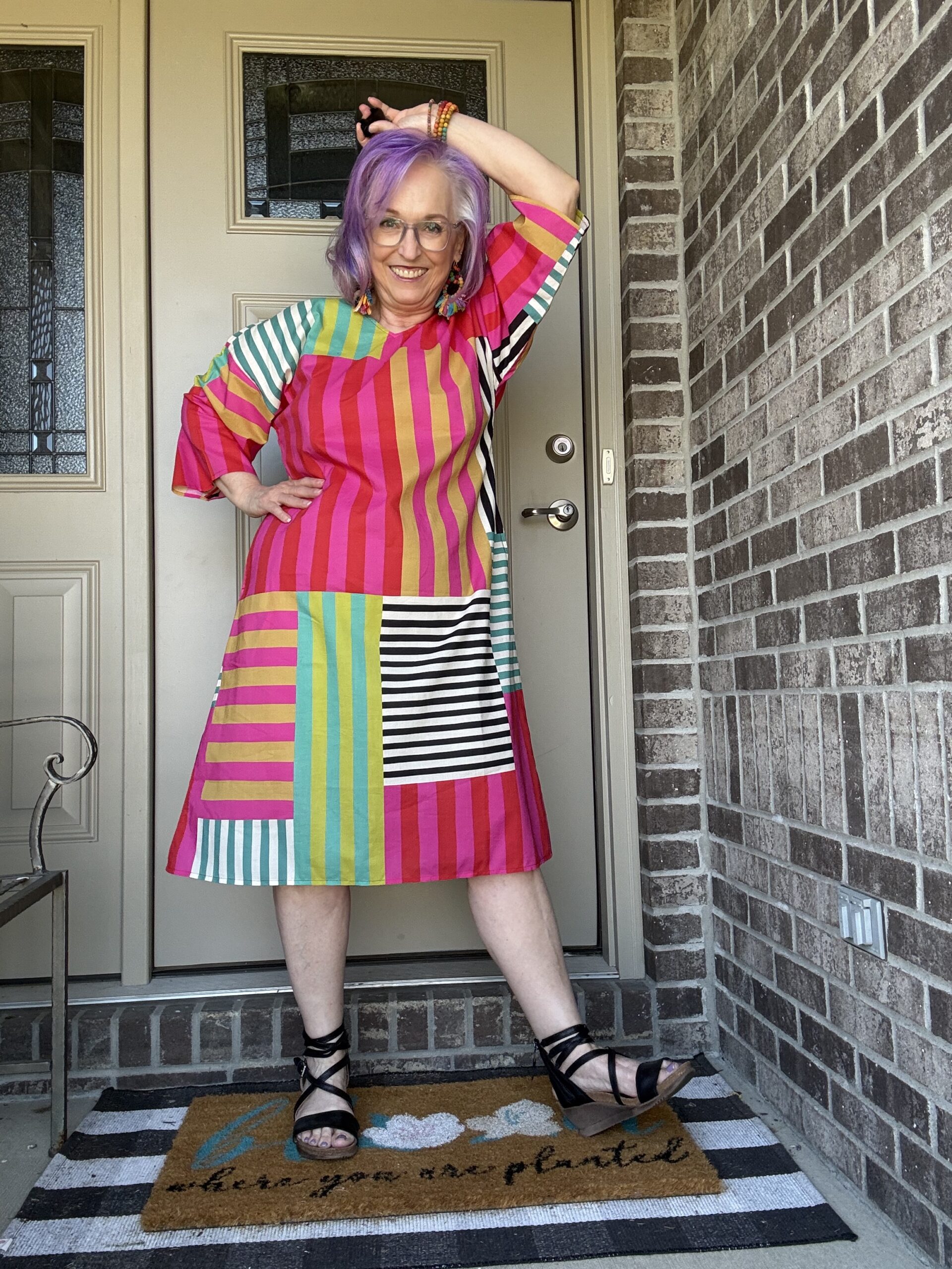 Woman with purple hair posing at a front door, wearing a bright, multicolored striped dress and black strappy sandals.