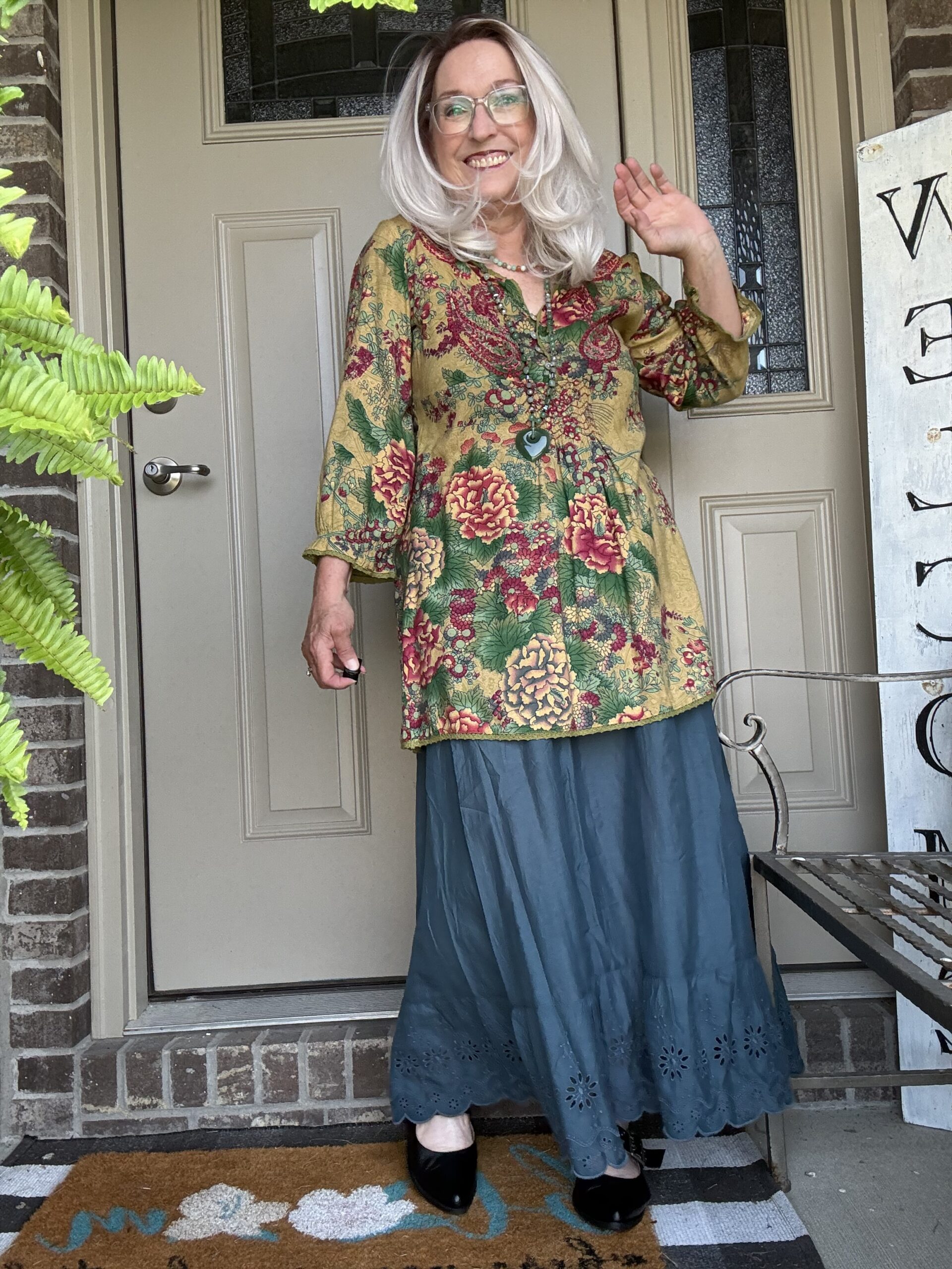 Smiling woman wearing a gold floral tunic and teal skirt stands on a front porch at a doorway, waving with dark shoes visible.