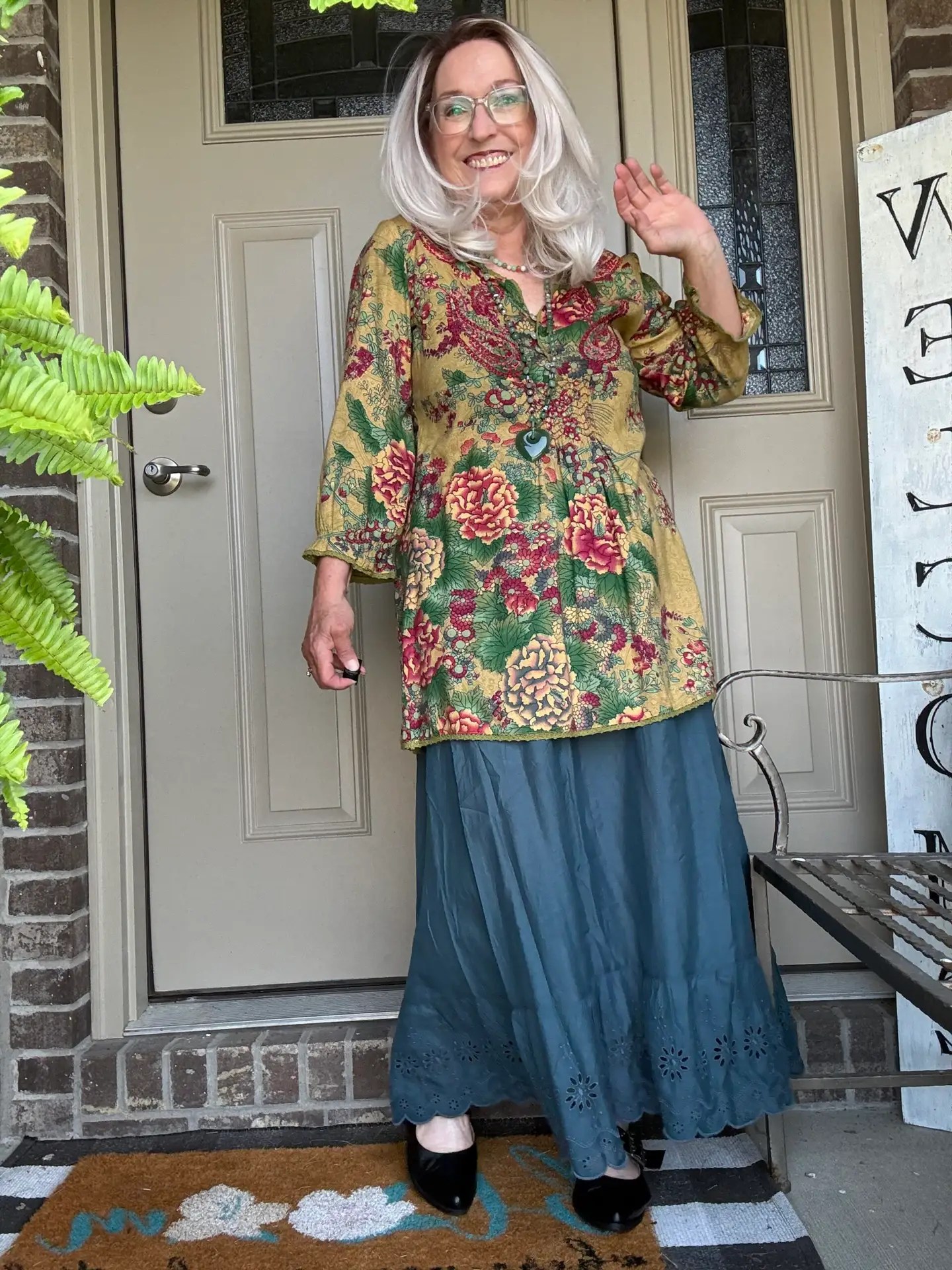 Smiling woman wearing a gold floral tunic and teal skirt stands on a front porch at a doorway, waving with dark shoes visible.