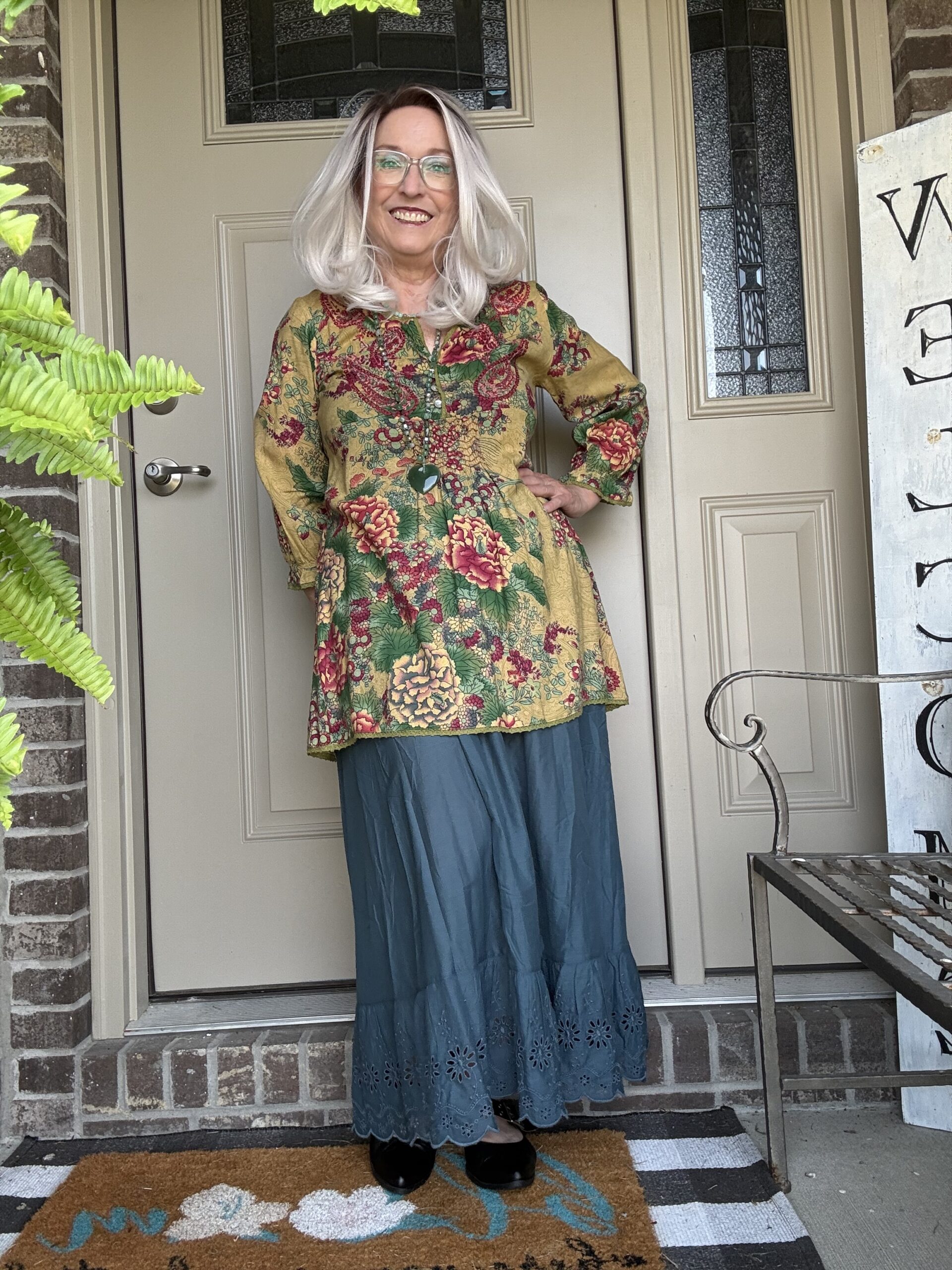 Smiling woman with silver hair and glasses standing on a porch in a mustard-yellow floral tunic and teal skirt, posing at the door.