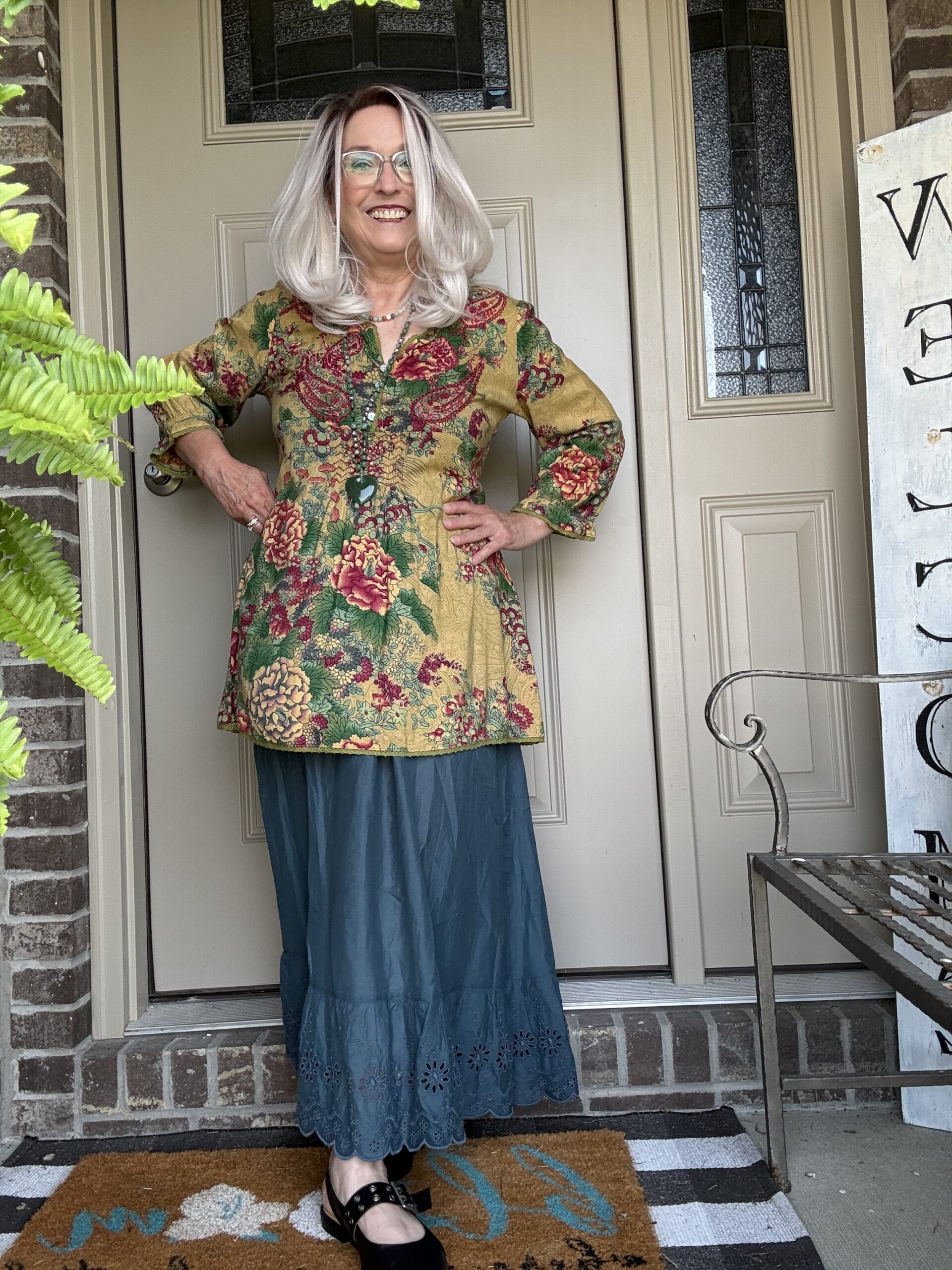 Smiling woman with silver hair and glasses stands on a porch in a mustard floral tunic and teal skirt, hands on hips.