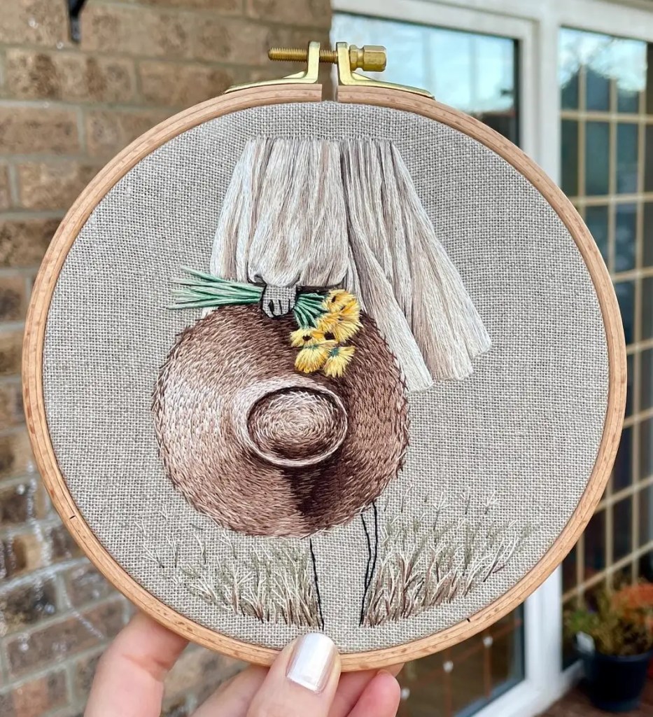 Decorative embroidery in an embroidery hoop: a brown hat with a white fringe and yellow flowers, set against a linen background with grass at the bottom.