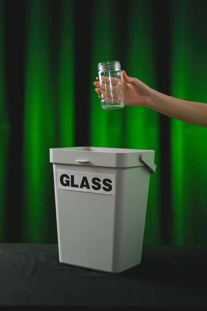 Hand holding a glass jar over a white bin labeled 'GLASS' with green spotlight backdrop.