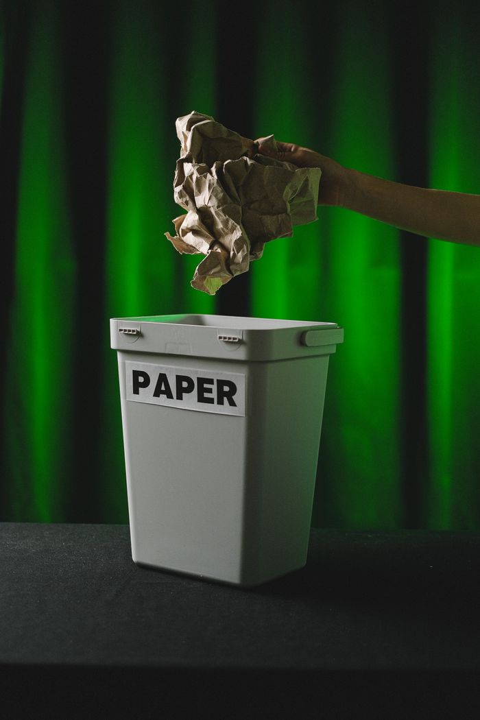 A hand discards crumpled paper into a labeled recycling bin against a green background.