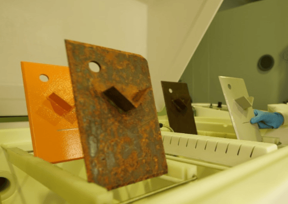 The image shows coated metal test panels, a salt spray corrosion chamber, and laboratory inspection equipment evaluating coating performance in accordance with ASTM D1654.