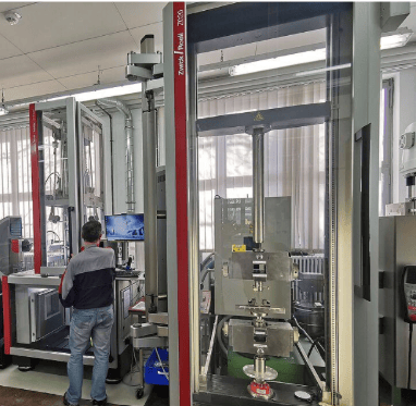 The image shows a universal testing machine measuring the ultimate tensile strength of a bolted longitudinal lapped seam in a deep corrugated structural steel plate during ASTM A1108.