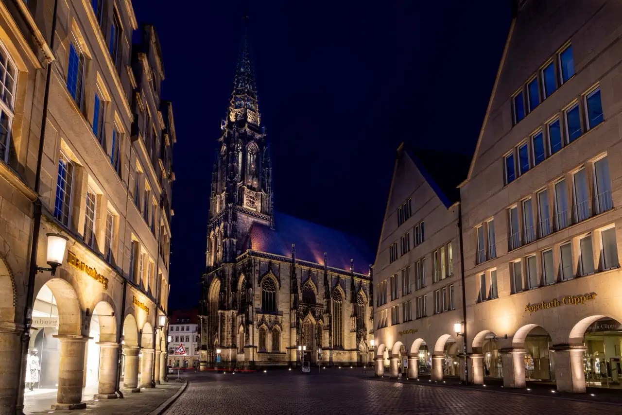 Ein nächtlicher Blick auf eine gotische Kathedrale mit einem hohen Turm neben modernen Gebäuden mit beleuchteten Schaufenstern auf einer Kopfsteinpflasterstraße.