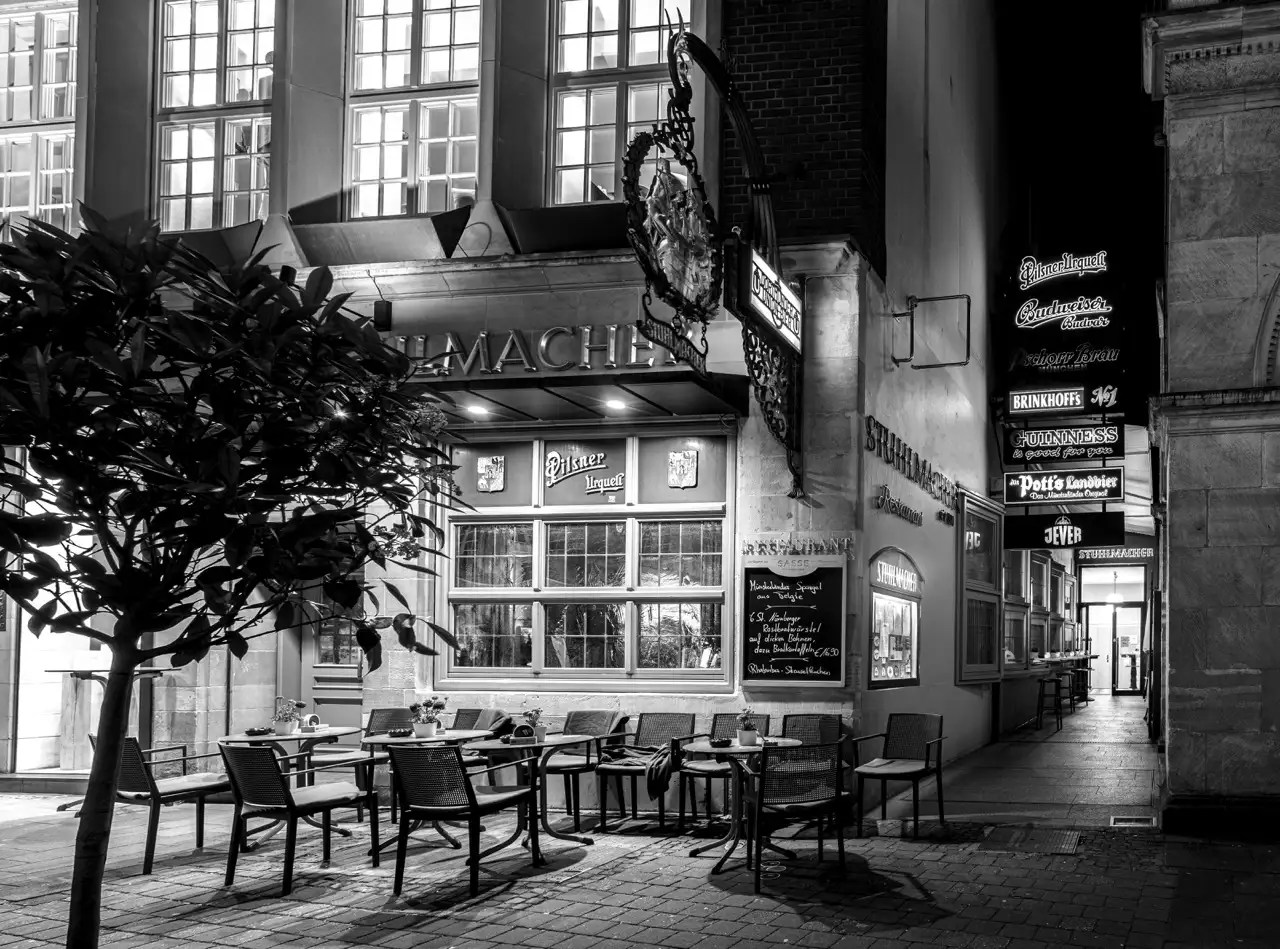 Schwarz-Weiß-Foto eines Sitzplatzes im Freien eines Restaurants mit einer Leuchtreklame, neben einem Fußgängerweg, der von verschnörkelten Werbeschildern für verschiedene Biermarken gesäumt ist.