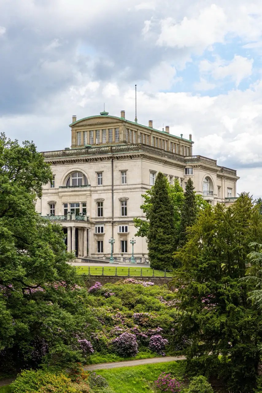 Ein großes, historisches Gebäude, umgeben von üppigem Grün und blühenden Sträuchern unter einem bewölkten Himmel.