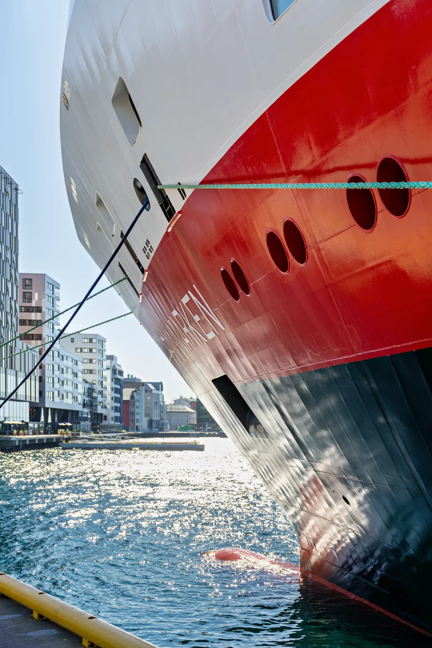 Bug eines großen Schiffes mit rotem und weißem Rumpf, vertäut im Hafen vor moderner Stadt mit Hochhäusern