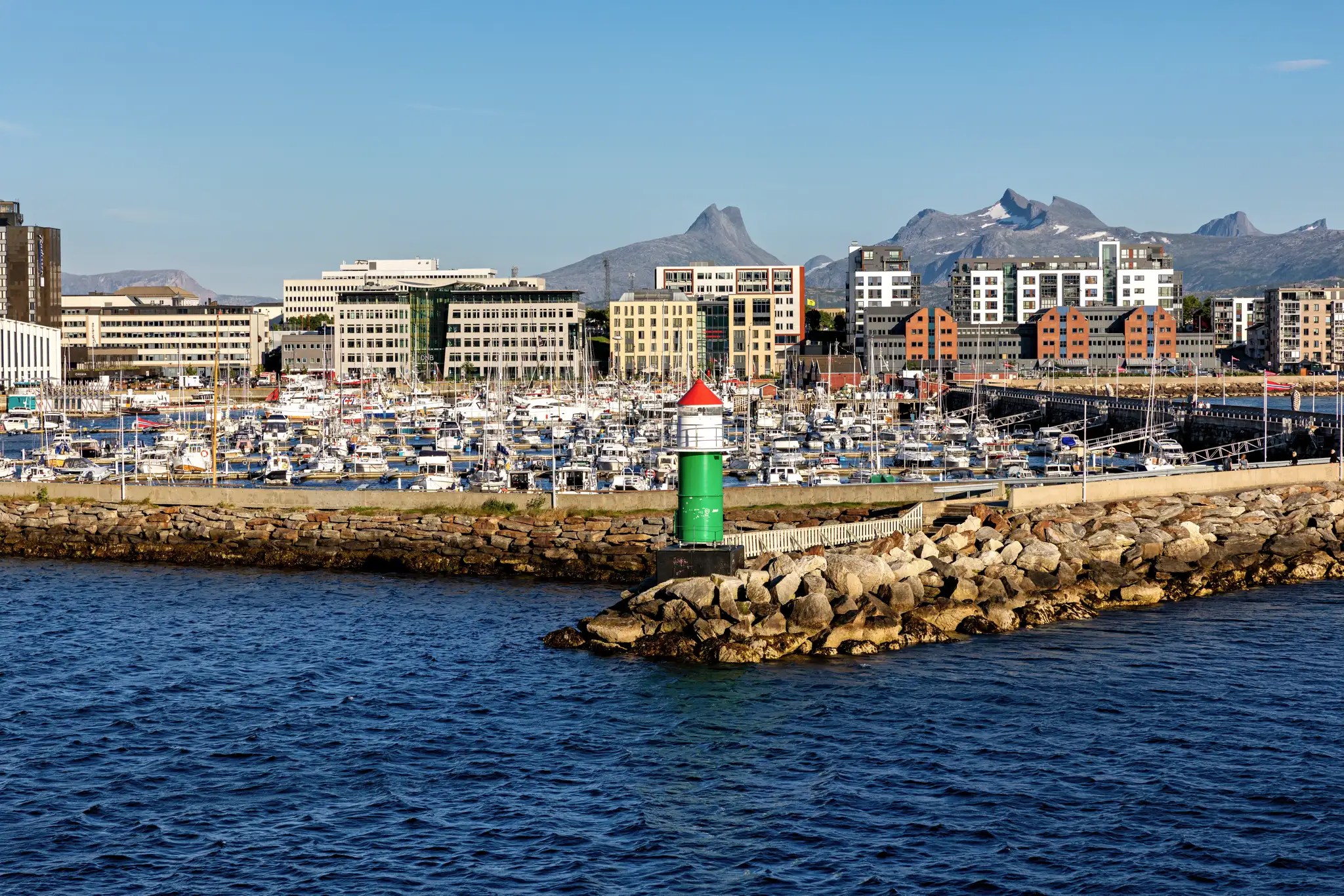 Grüner und weißer Leuchtturm mit rotem Dach am Hafen von Bodø, Norwegen, mit Booten und modernen Gebäuden vor Bergen im Hintergrund