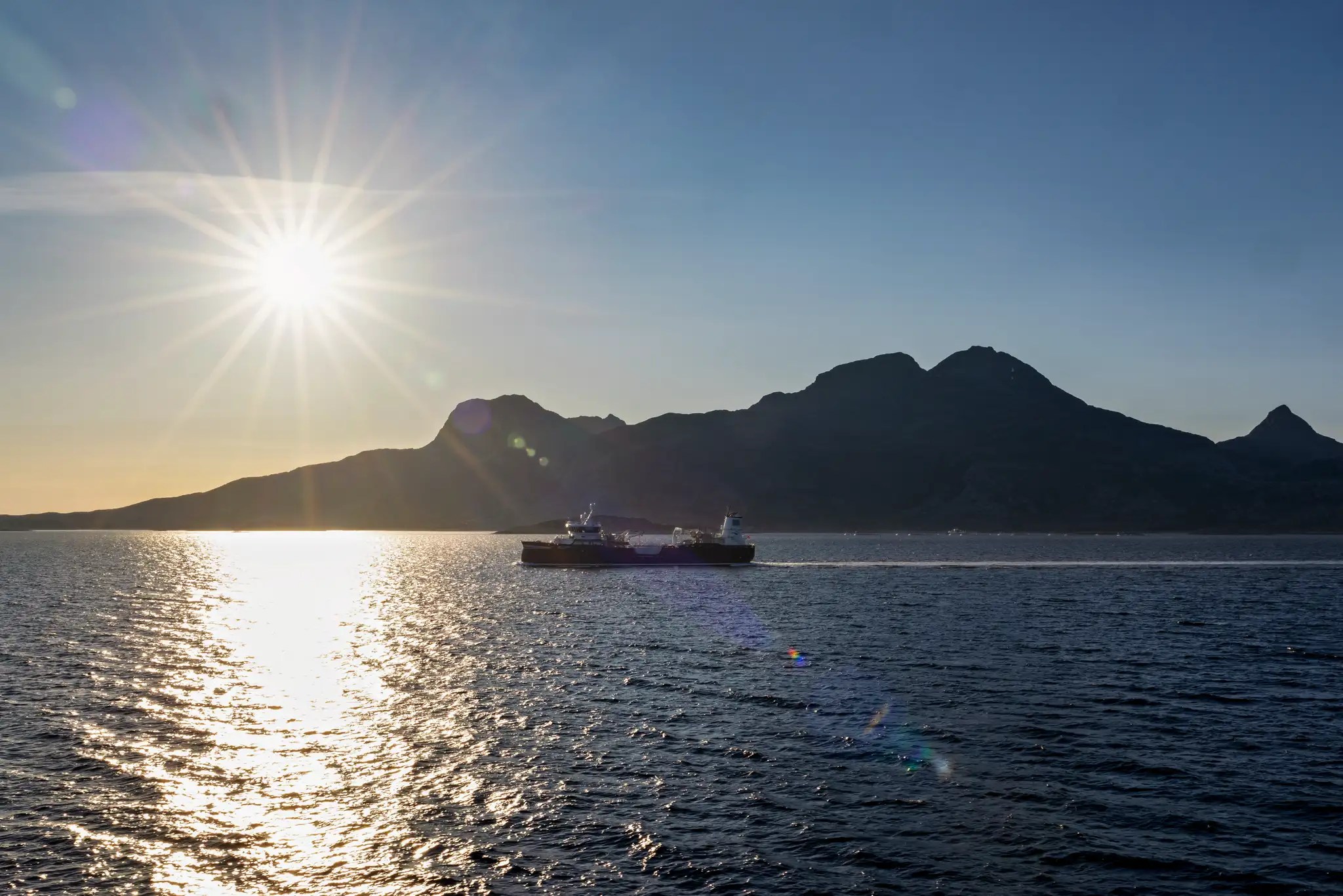 Sonnenuntergang über einem ruhigen Meer mit einem Frachtschiff vor einer Bergsilhouette