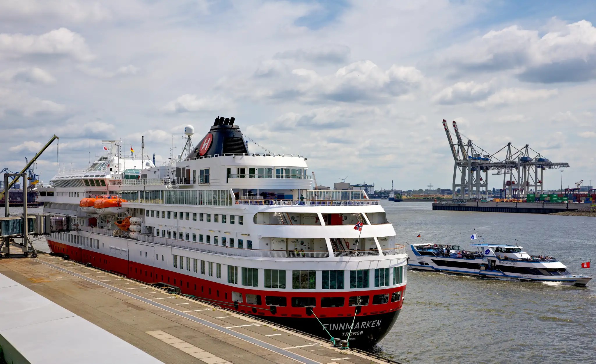 Das Kreuzfahrtschiff Finnmarken der Hurtigruten liegt am Kai eines Hafens, im Hintergrund Containerkräne und ein kleineres Boot auf dem Wasser