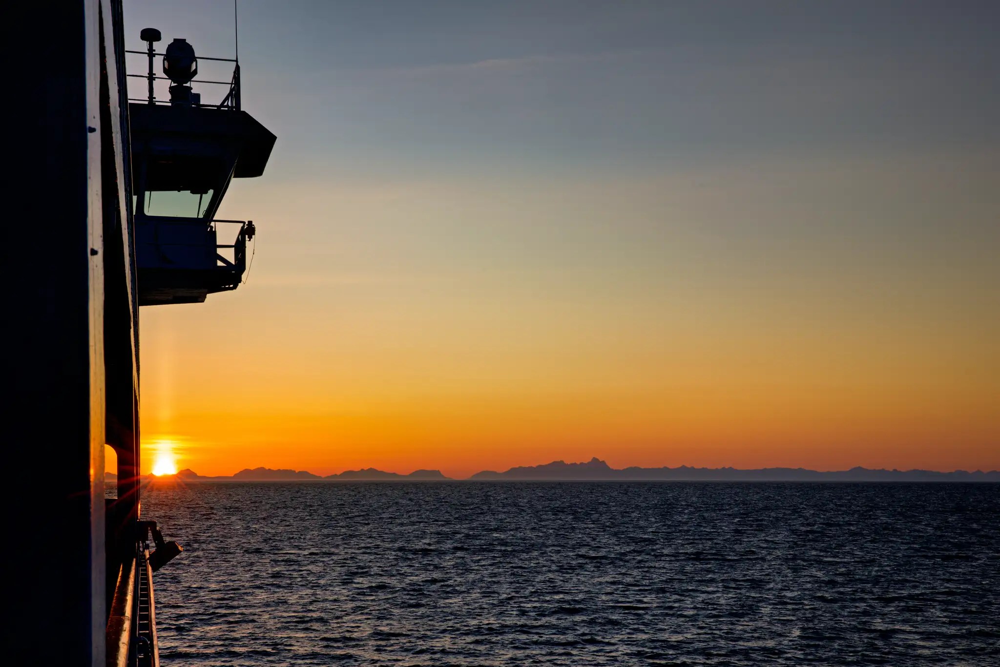 Sonnenuntergang über dem Meer mit Silhouetten von Bergen, gesehen von der Brücke eines Schiffes