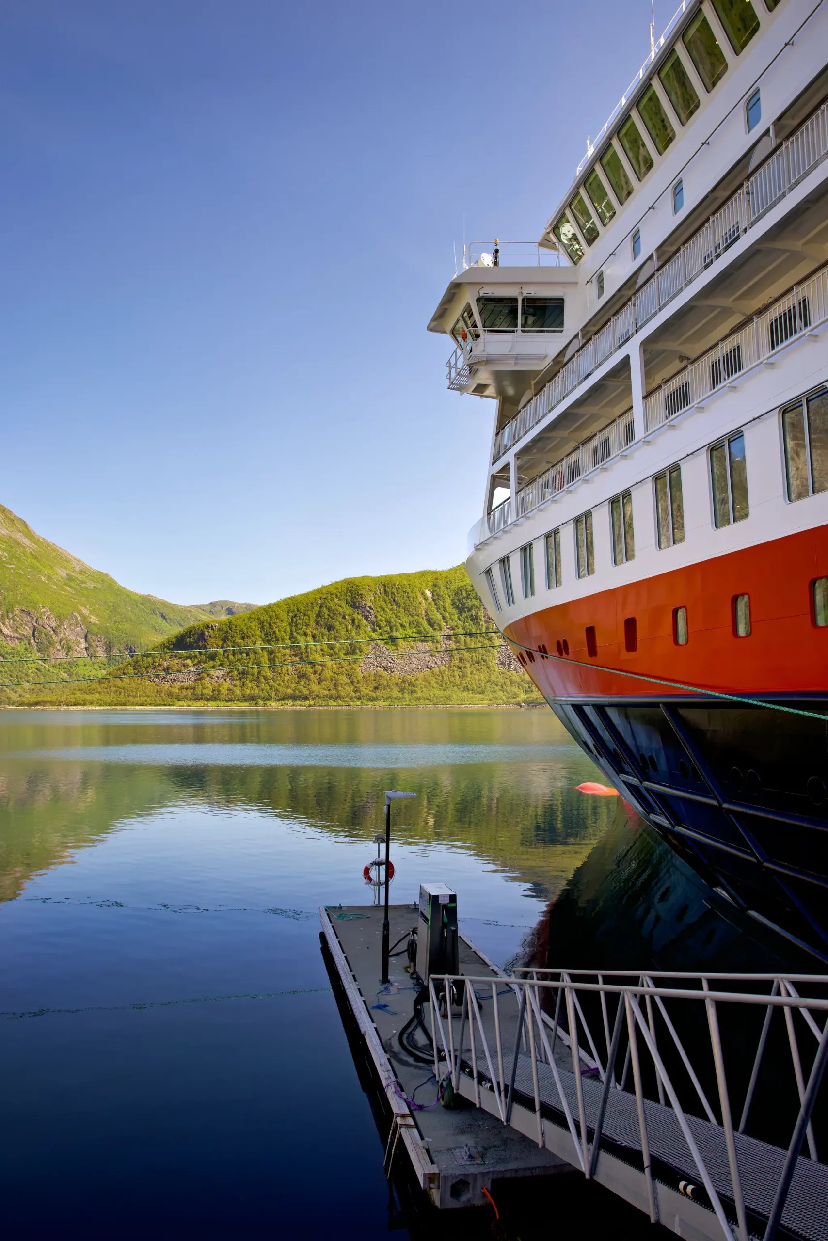 Vorderer Teil eines Hurtigruten-Kreuzfahrtschiffs am Kai in einem ruhigen Fjord mit grünen Bergen im Hintergrund