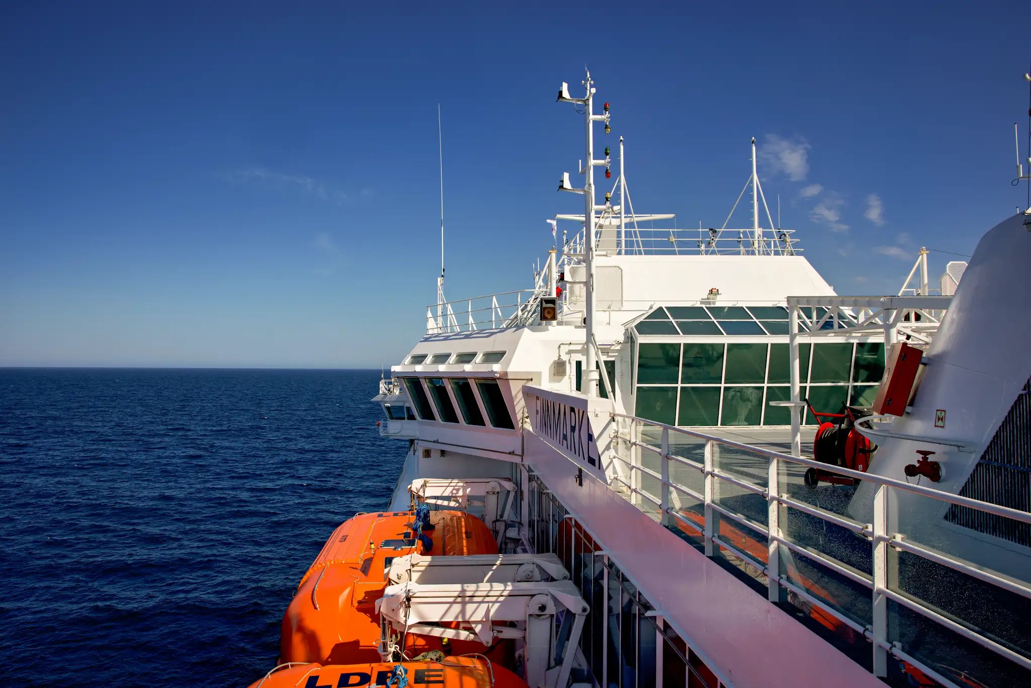 Blick auf das Deck eines Hurtigruten-Kreuzfahrtschiffs mit Rettungsbooten und offenem Meer unter klarem Himmel