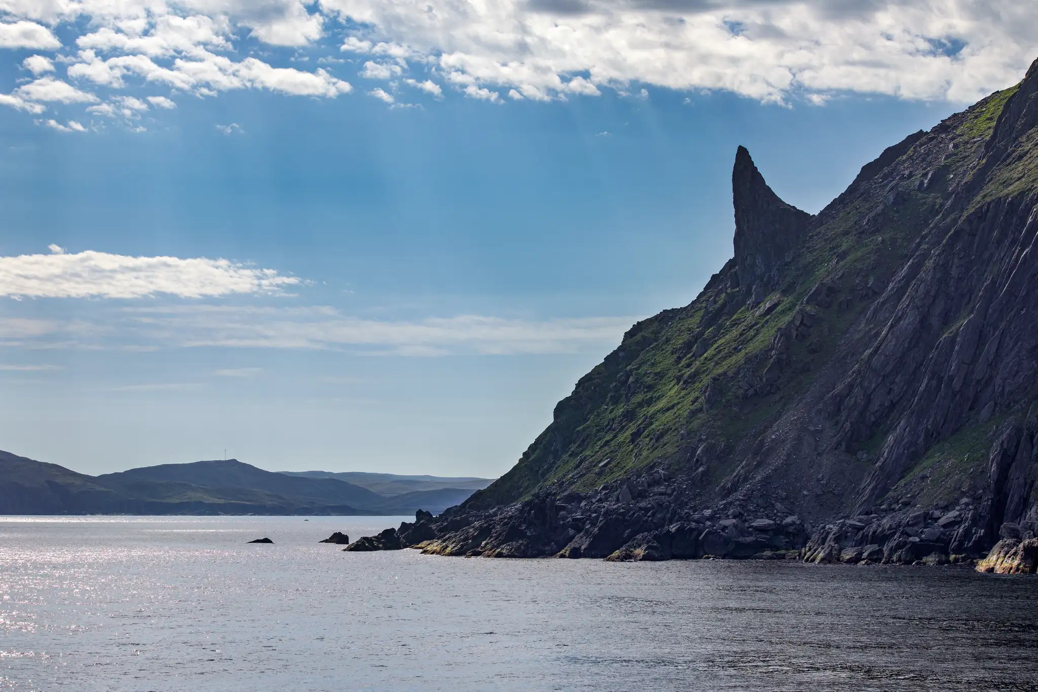 Felsige Küste mit steilem, spitz zulaufendem Berg unter bewölktem Himmel am Meer