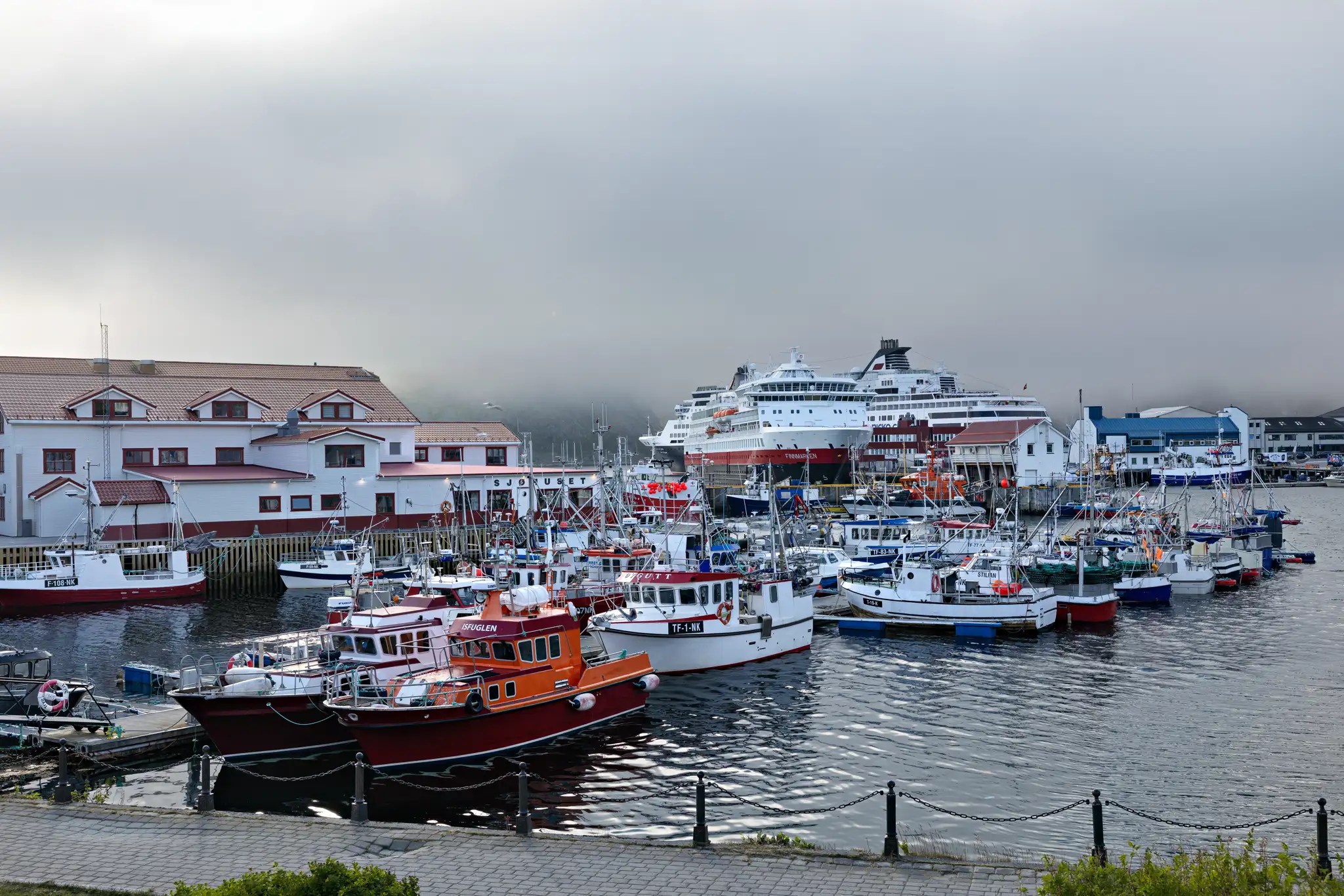 Hafen mit mehreren kleinen Booten und einem großen Hurtigruten-Kreuzfahrtschiff im Hintergrund unter bewölktem Himmel