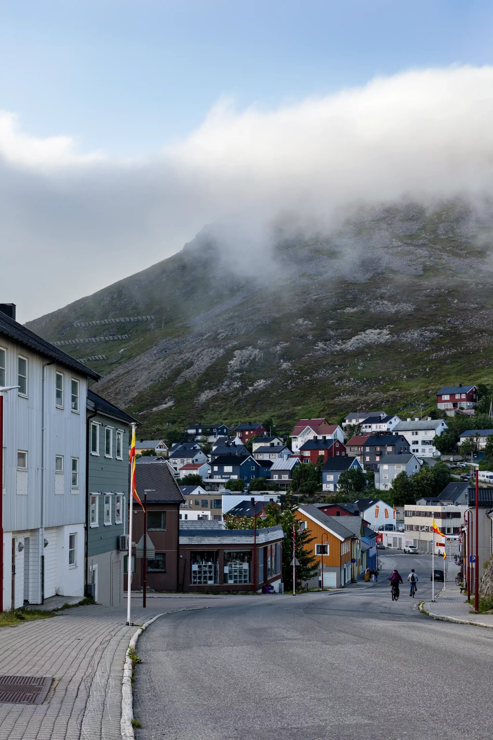 Straße mit bunten Häusern und zwei Radfahrern vor nebelverhülltem Berg in norwegischem Küstenort