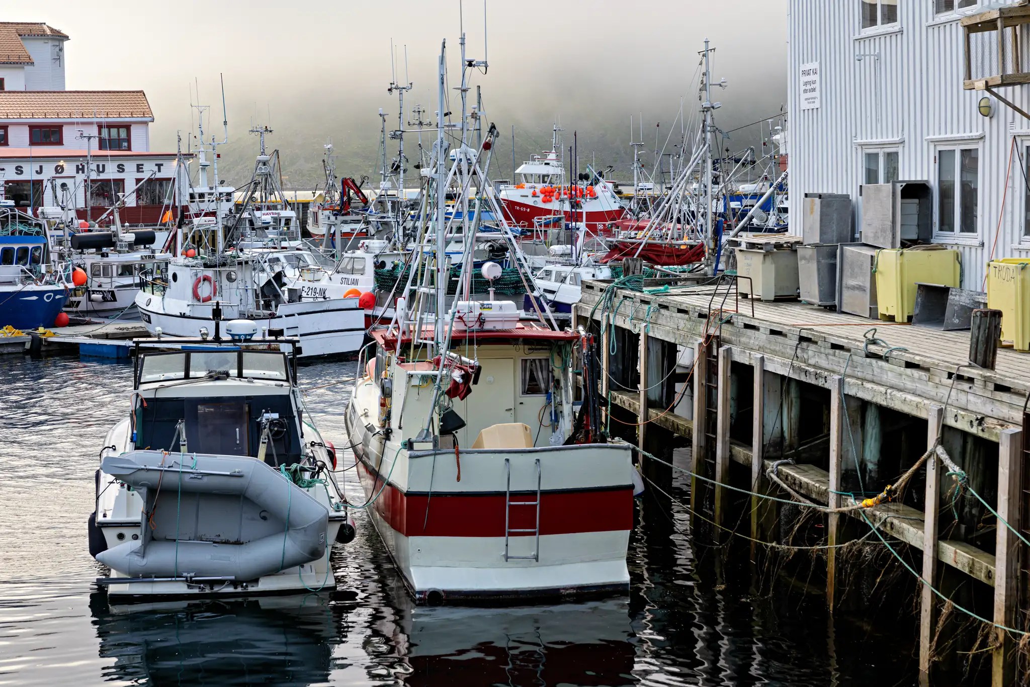 Mehrere Fischerboote und Schiffe liegen an einem Hafensteg, im Hintergrund ein Gebäude mit der Aufschrift 'Sjøhuset'
