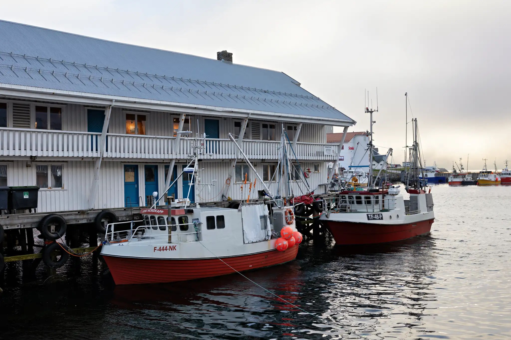 Zwei rote und weiße Fischerboote am Kai vor einem weißen Holzgebäude mit blauen Türen im Hafen