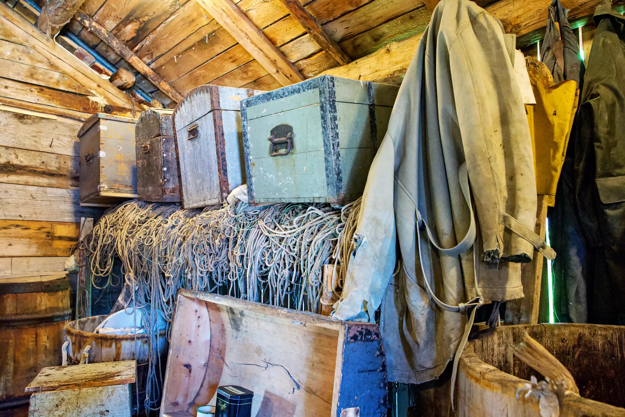 Alter Lagerraum mit Holzwänden, mehreren Stapeln von Seilen, Metallkisten und hängender Arbeitskleidung