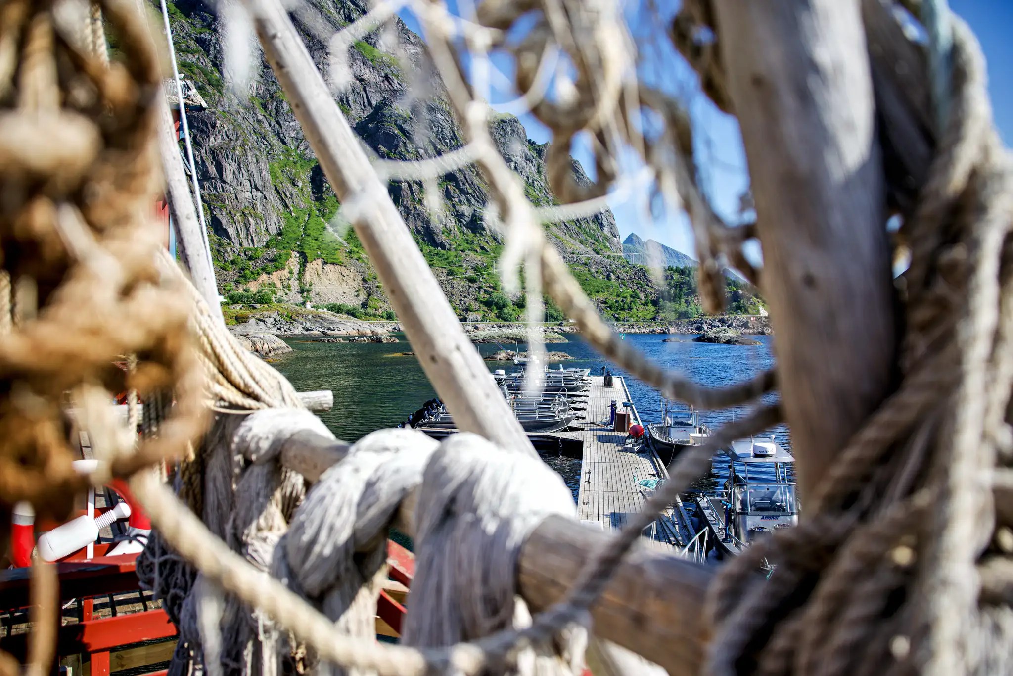 Blick durch ein Netz aus dicken Seilen auf einen Pier mit Booten und felsige grüne Berge im Hintergrund