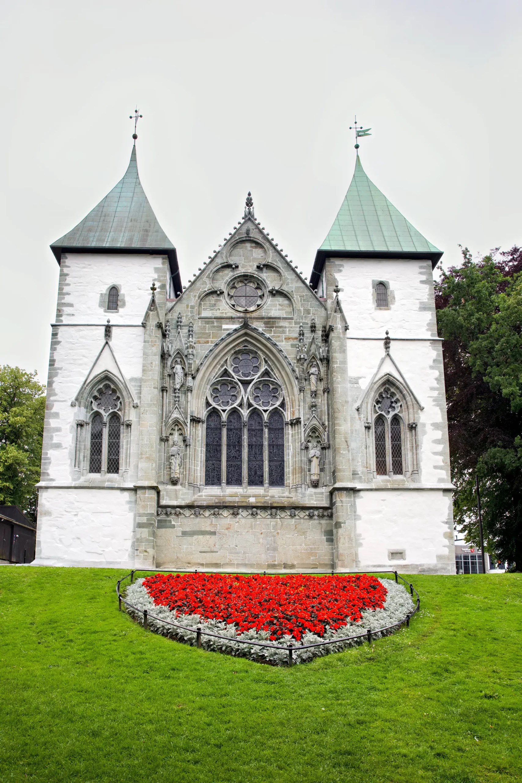 Gotische Kirche mit zwei Türmen und einem dreieckigen Giebel, davor ein Blumenbeet mit roten Blumen auf grünem Rasen