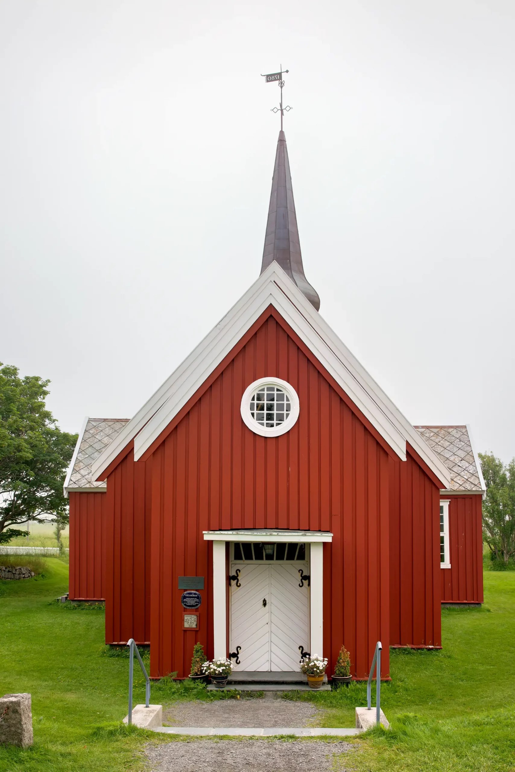 Rotes Holzkirchgebäude mit weißem Eingang und spitzem Turm auf grünem Rasen