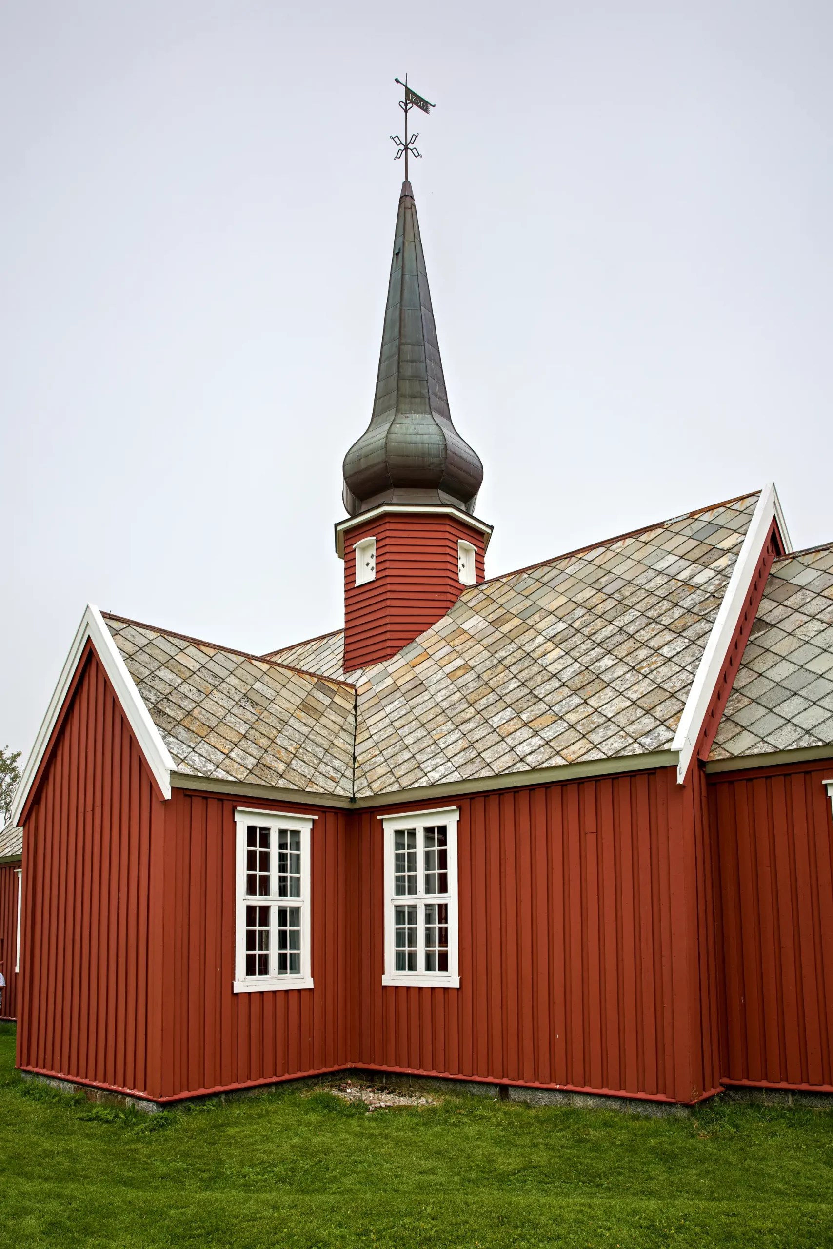 Rotes Holzgebäude mit spitzem Turm und Wetterfahne, schräges Schindeldach, grüne Wiese im Vordergrund
