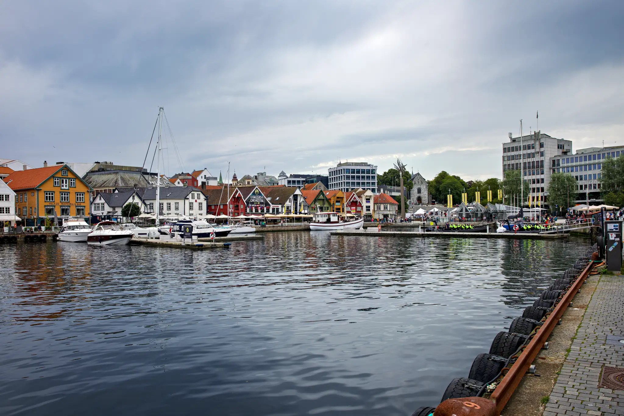 Hafen mit mehreren Segel- und Motorbooten vor einer Reihe bunter Häuser und moderner Gebäude unter bewölktem Himmel