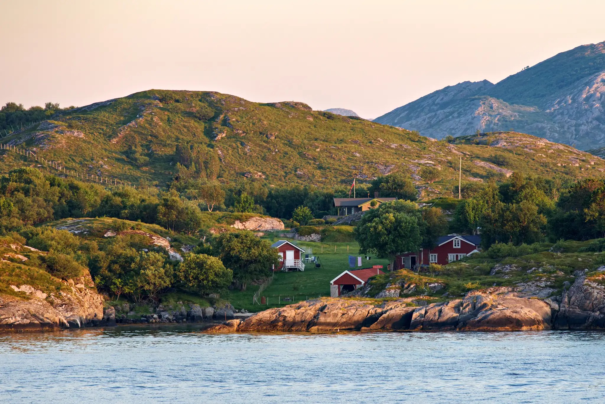Zwei rote Holzhäuser am Ufer eines Fjords, umgeben von grünen Hügeln und Bergen im Hintergrund