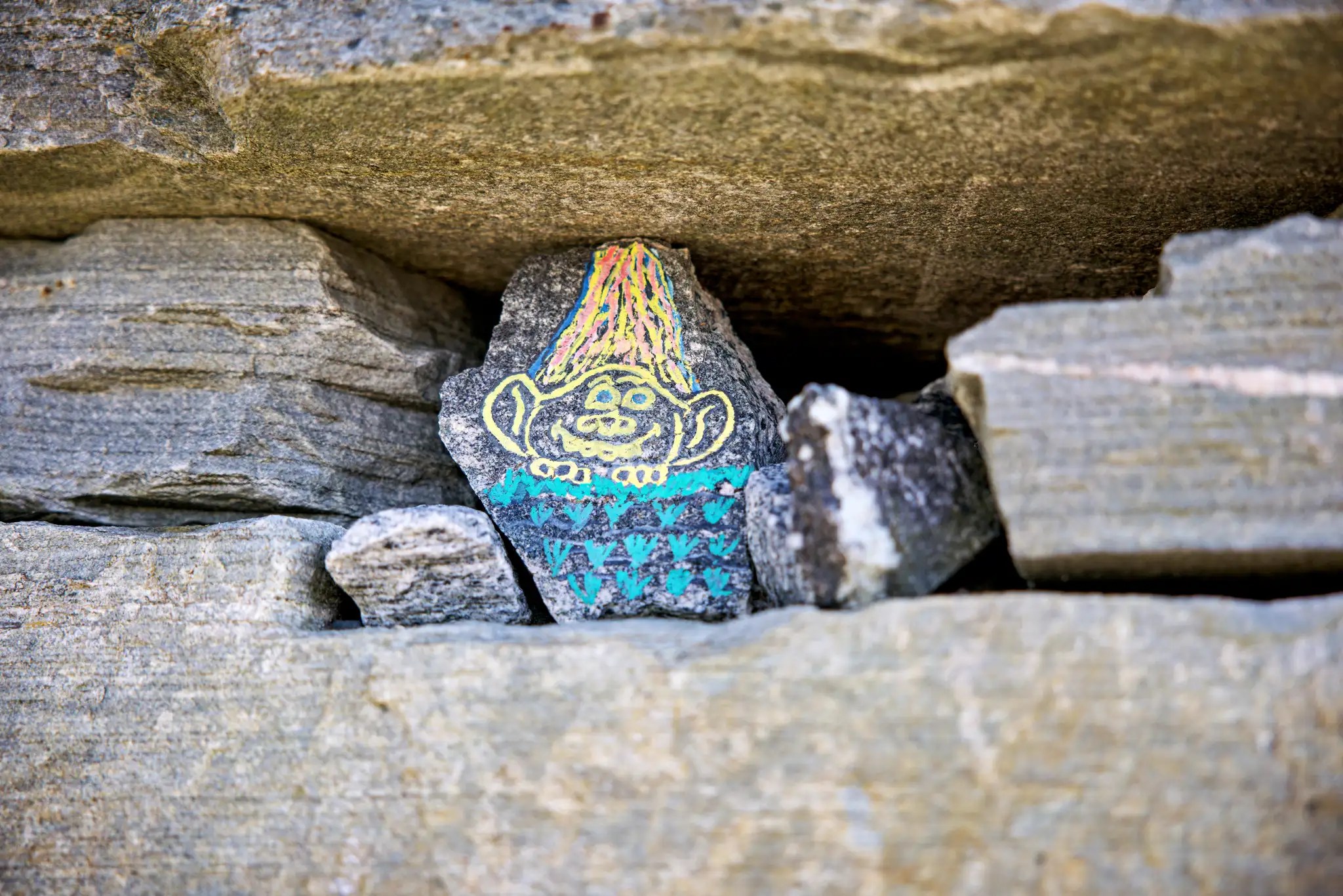 Kleiner Stein mit bunter Trollzeichnung, platziert zwischen größeren grauen Steinen