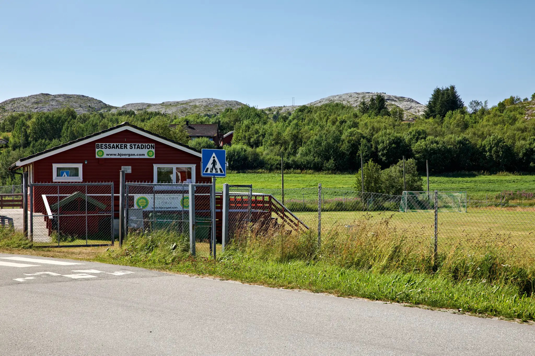 Rotes Gebäude mit Schild 'Bessaker Stadion' neben einem Fußballfeld und grünen Hügeln im Hintergrund
