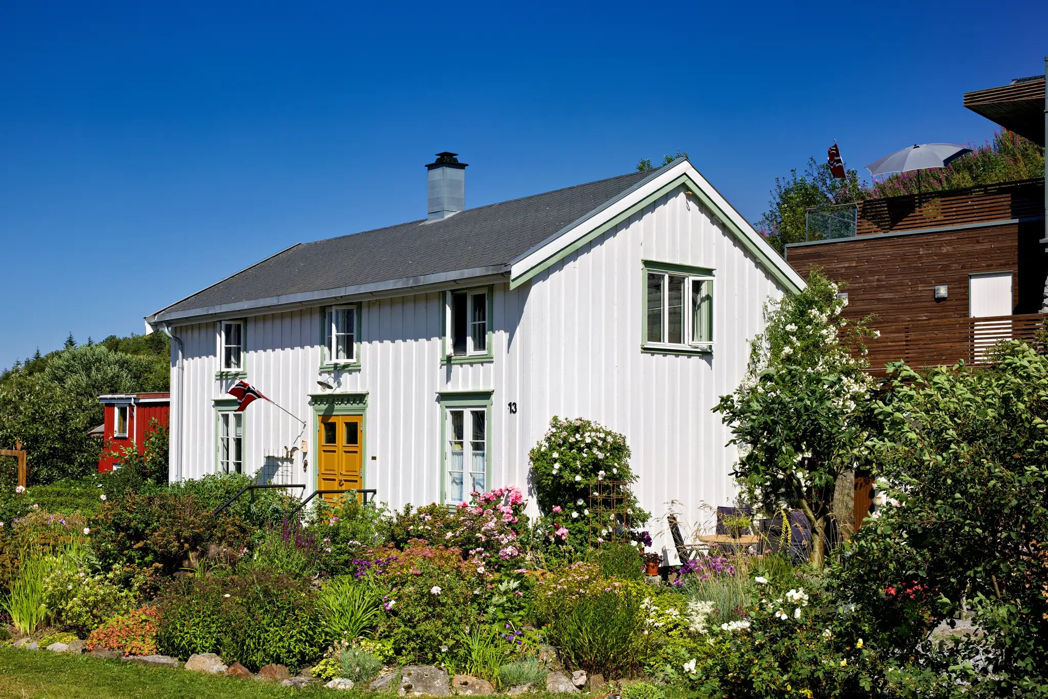 Weißes zweistöckiges Haus mit gelber Tür, umgeben von buntem Garten und blühenden Sträuchern unter blauem Himmel