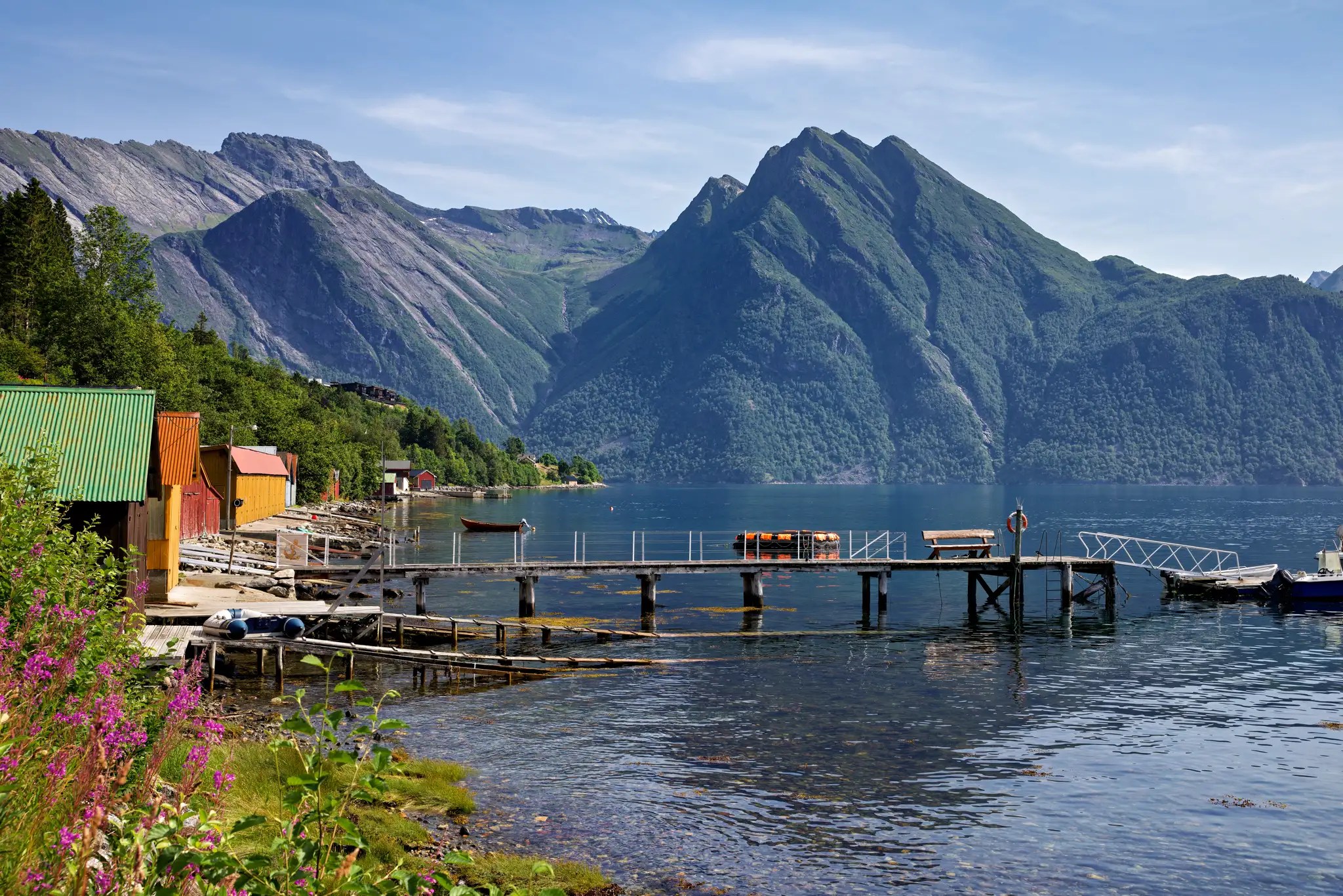 Fjord mit steilen Bergen im Hintergrund, Holzsteg und bunte Häuser am Ufer, ruhiges Wasser unter blauem Himmel