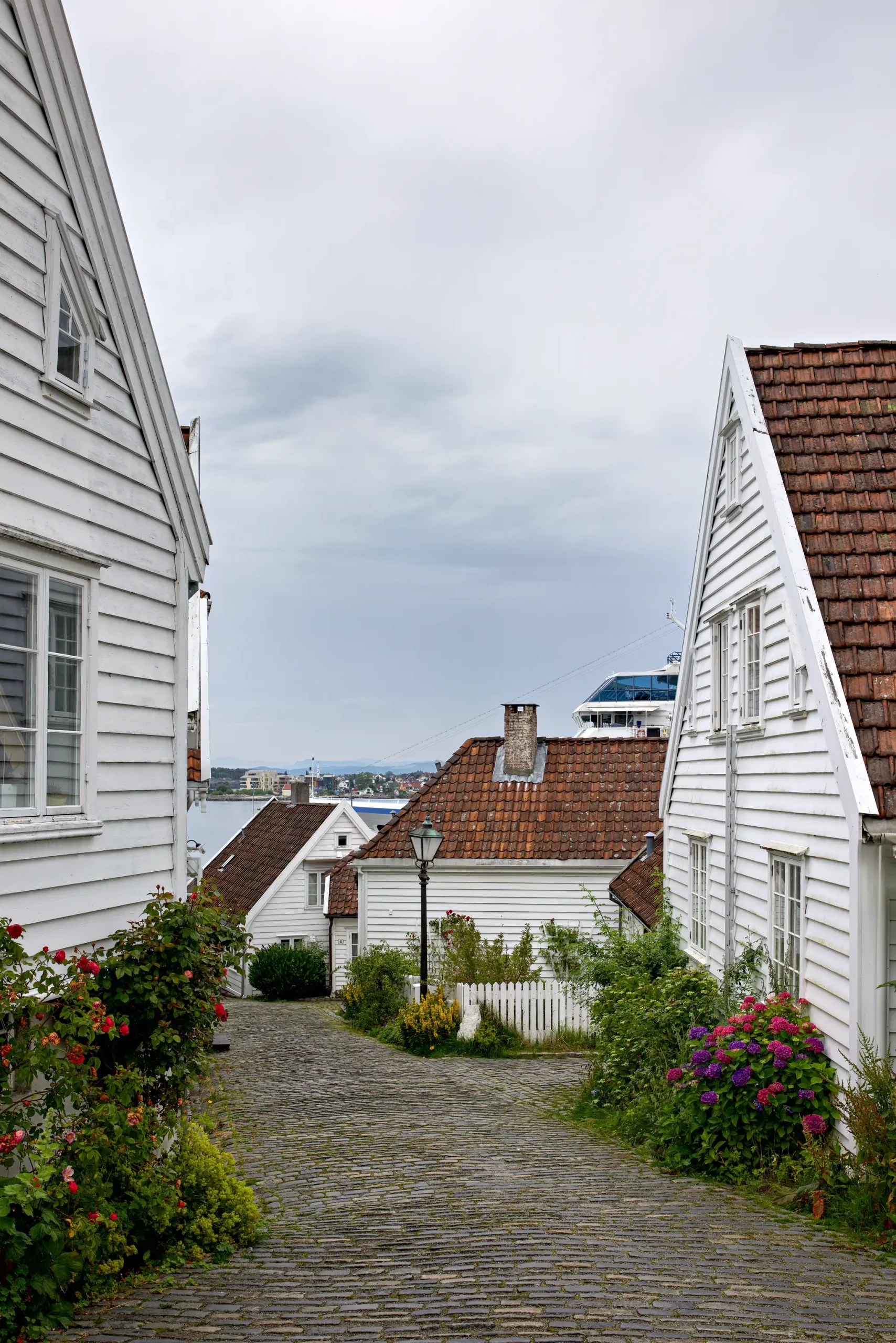 Kopfsteinpflasterstraße zwischen weißen Holzhäusern mit roten Dächern und Blumen, im Hintergrund Meer und Schiff