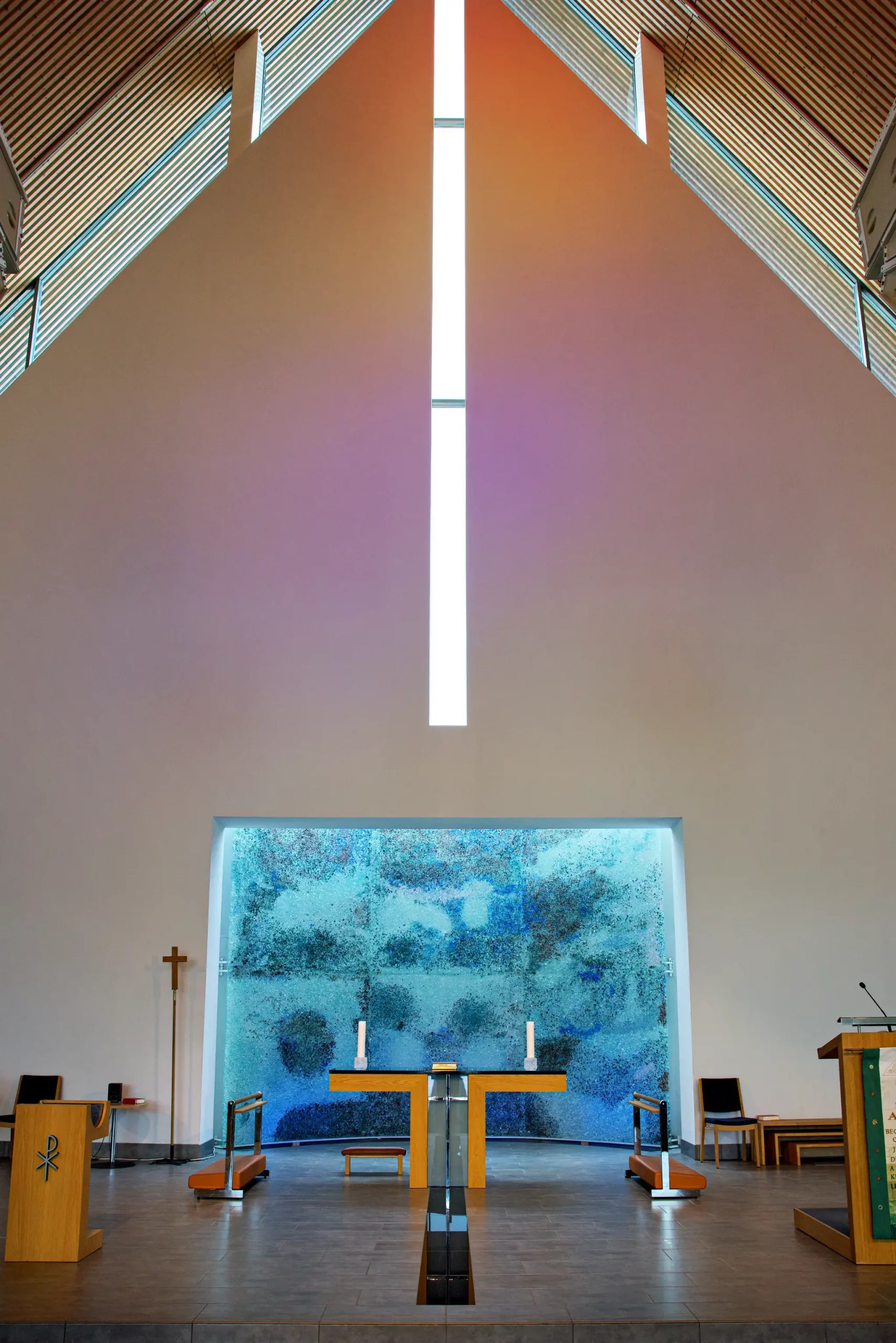 Innenraum einer modernen Kirche mit hohem, spitz zulaufendem Dach, zentralem Altar und großem Fenster mit abstraktem Muster