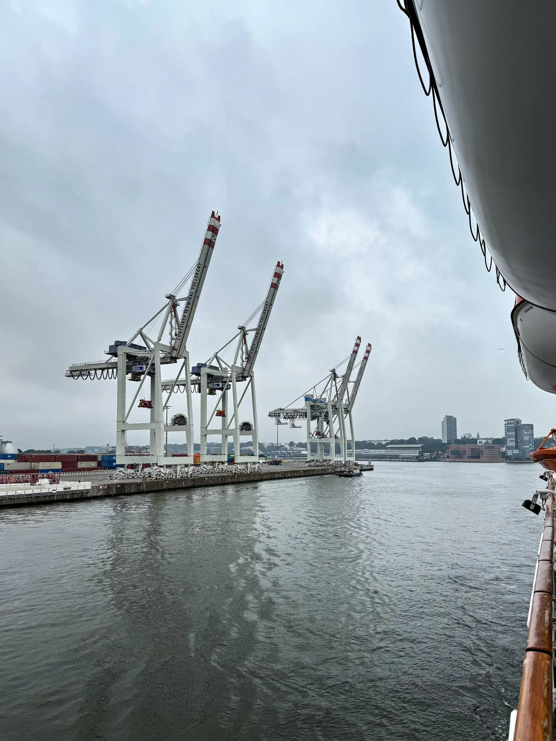 Blick von einem Schiff auf Hafenkräne und Container am Wasser unter bewölktem Himmel