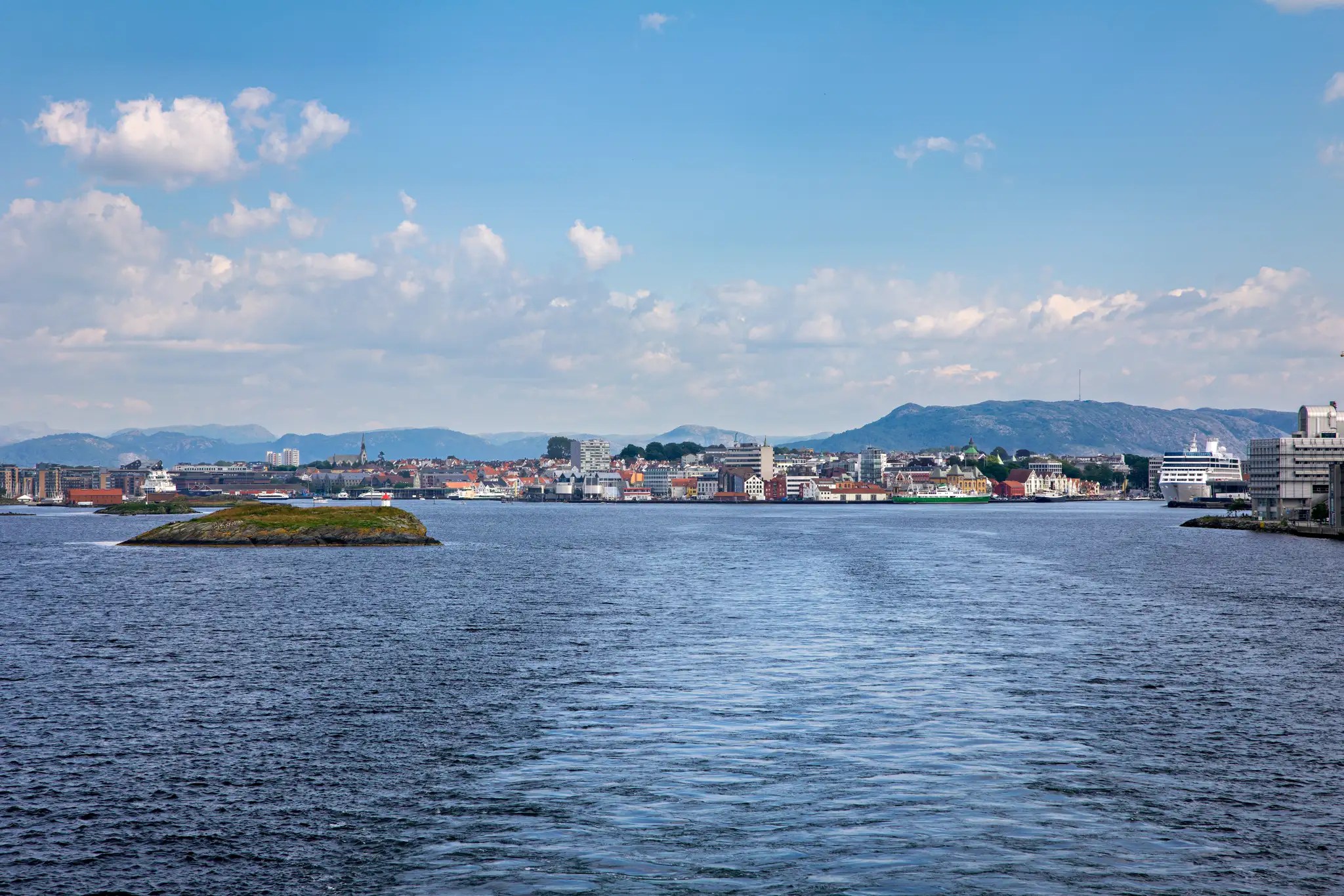 Blick auf eine Hafenstadt mit Bergen im Hintergrund und Wasser im Vordergrund unter blauem Himmel mit Wolken
