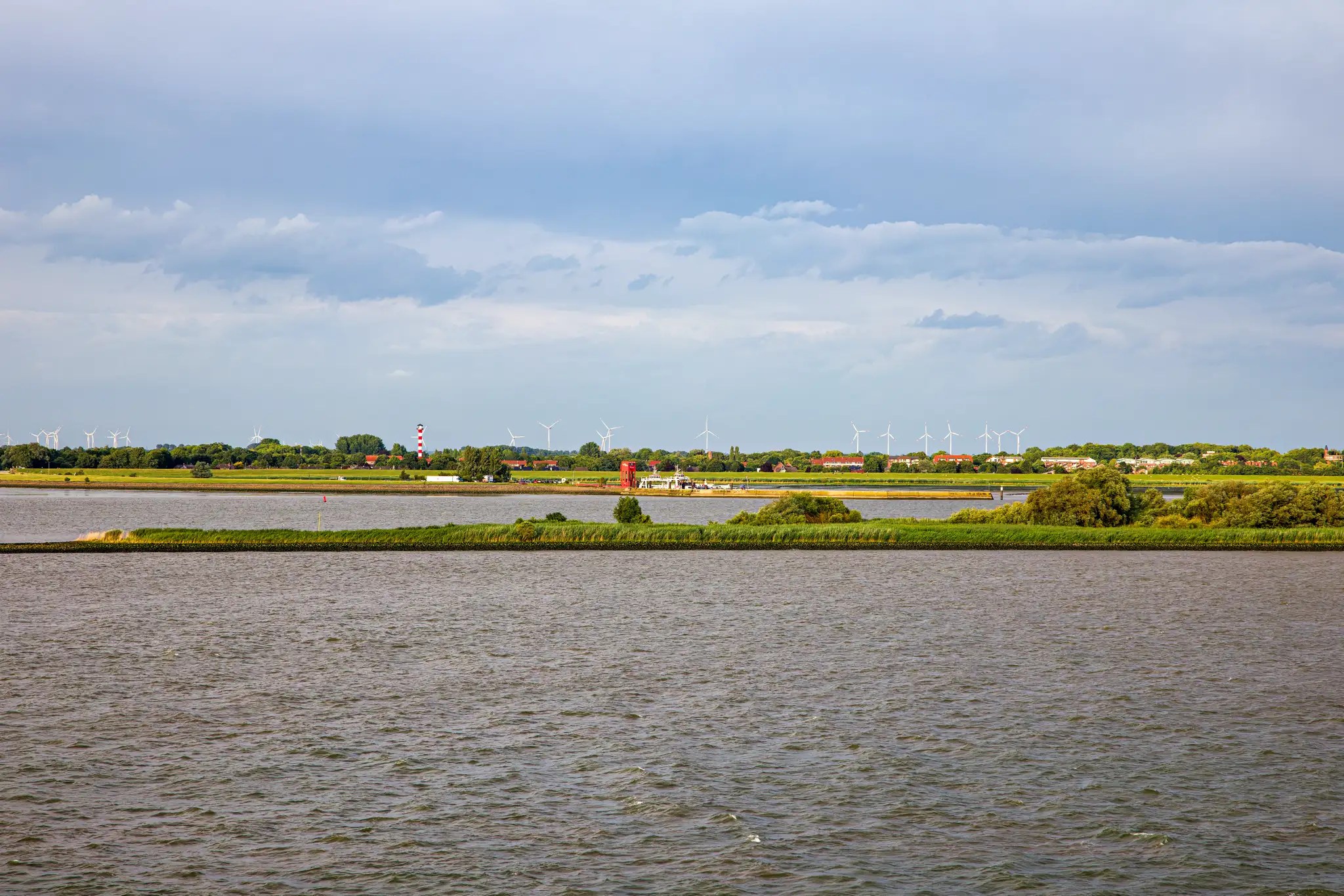 Flusslandschaft mit grünen Inseln und Windrädern am Horizont unter bewölktem Himmel