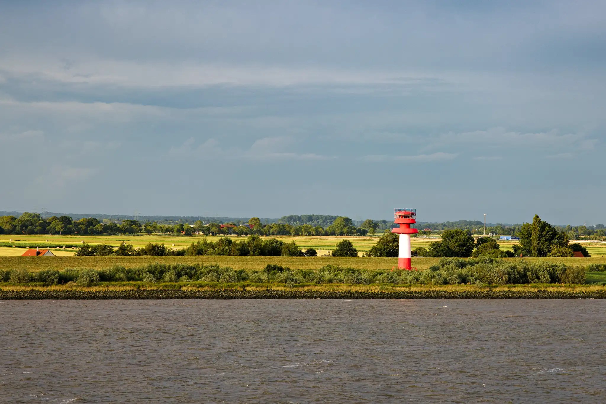Leuchtturm mit rotem und weißem Streifen an der Küste, dahinter grüne Felder und bewölkter Himmel
