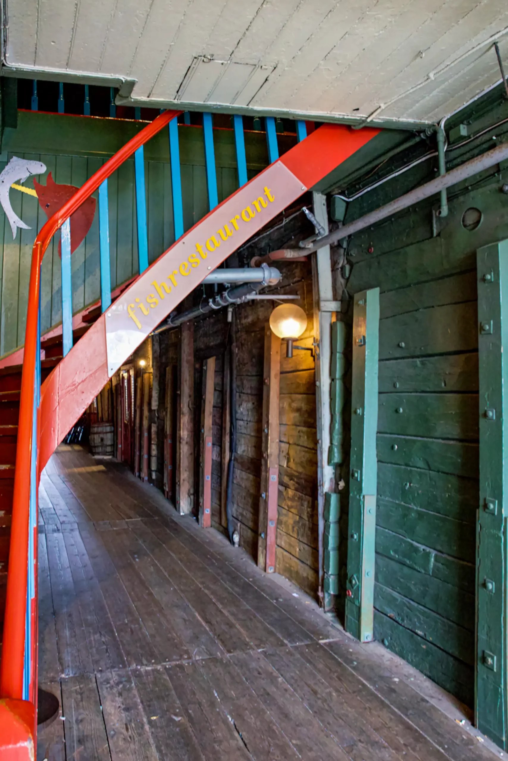 Holztreppe mit rotem Geländer und gelbem Schriftzug 'fishrestaurant' in einem historischen Lagerhaus mit grünen Holzwänden