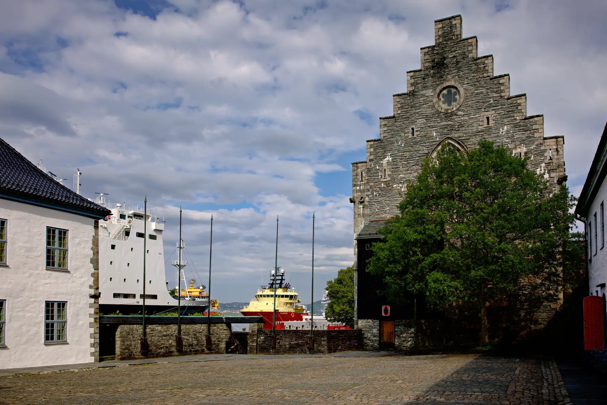 Historisches Gebäude mit Treppengiebel neben Bäumen, im Hintergrund zwei Hurtigruten-Schiffe im Hafen