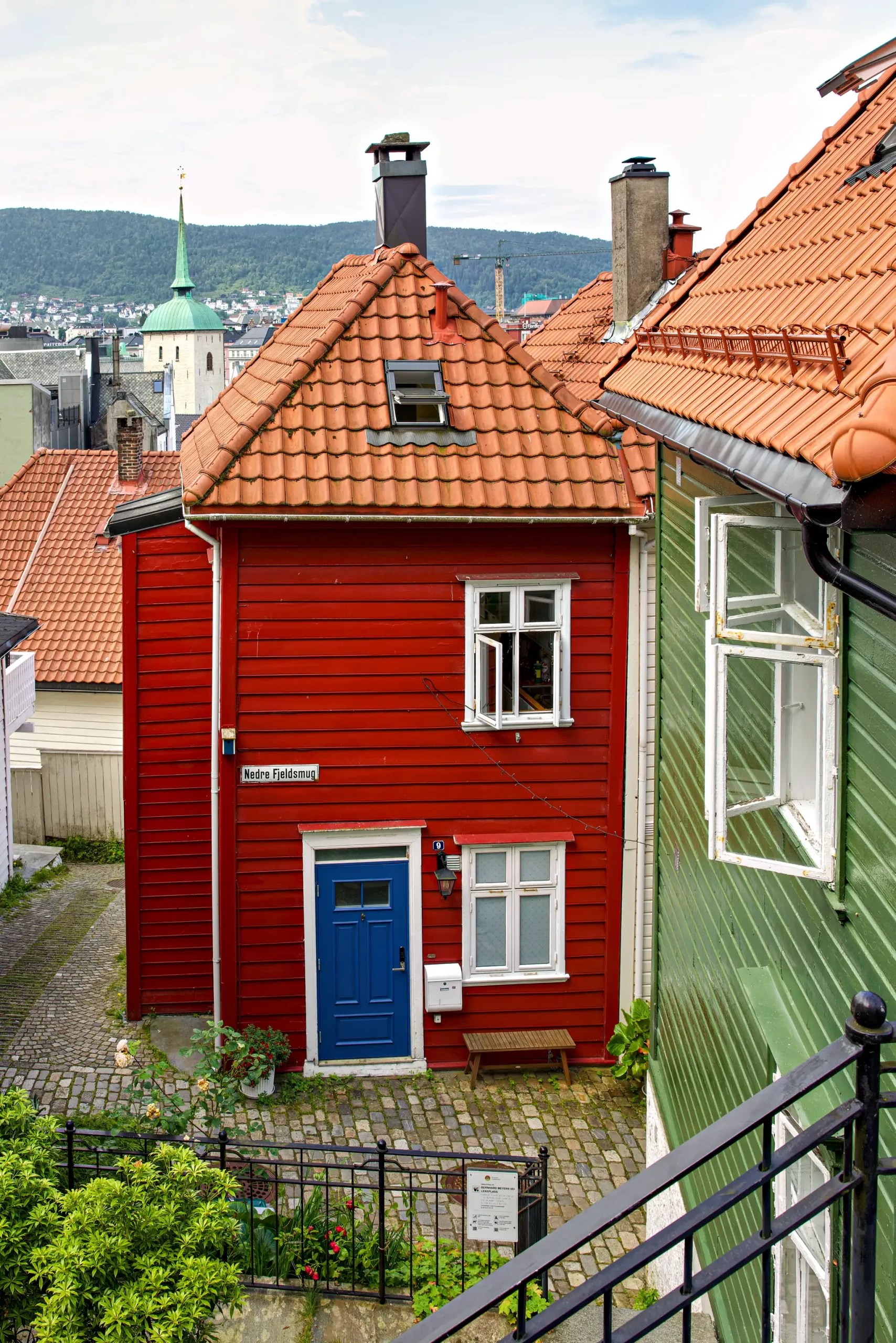 Rotes Holzhaus mit blauem Eingang und zwei Fenstern in einer historischen Straße, umgeben von grünen und roten Häusern mit roten Ziegeldächern